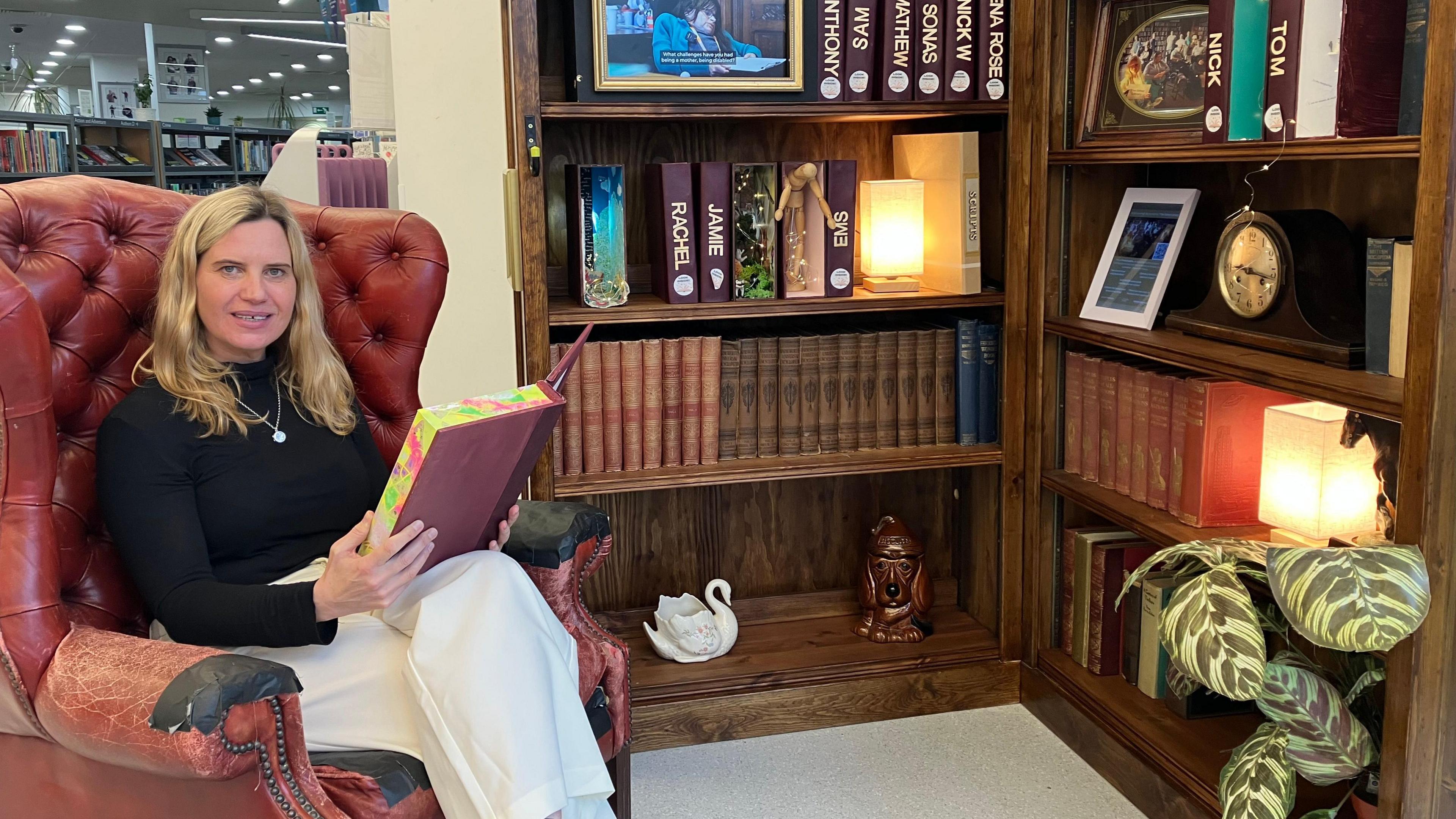 A picture of a blonde woman who is sitting on a red leather chair with a book in her hands. To her right is a bookcase. She is wearing a black top and has white trousers on.