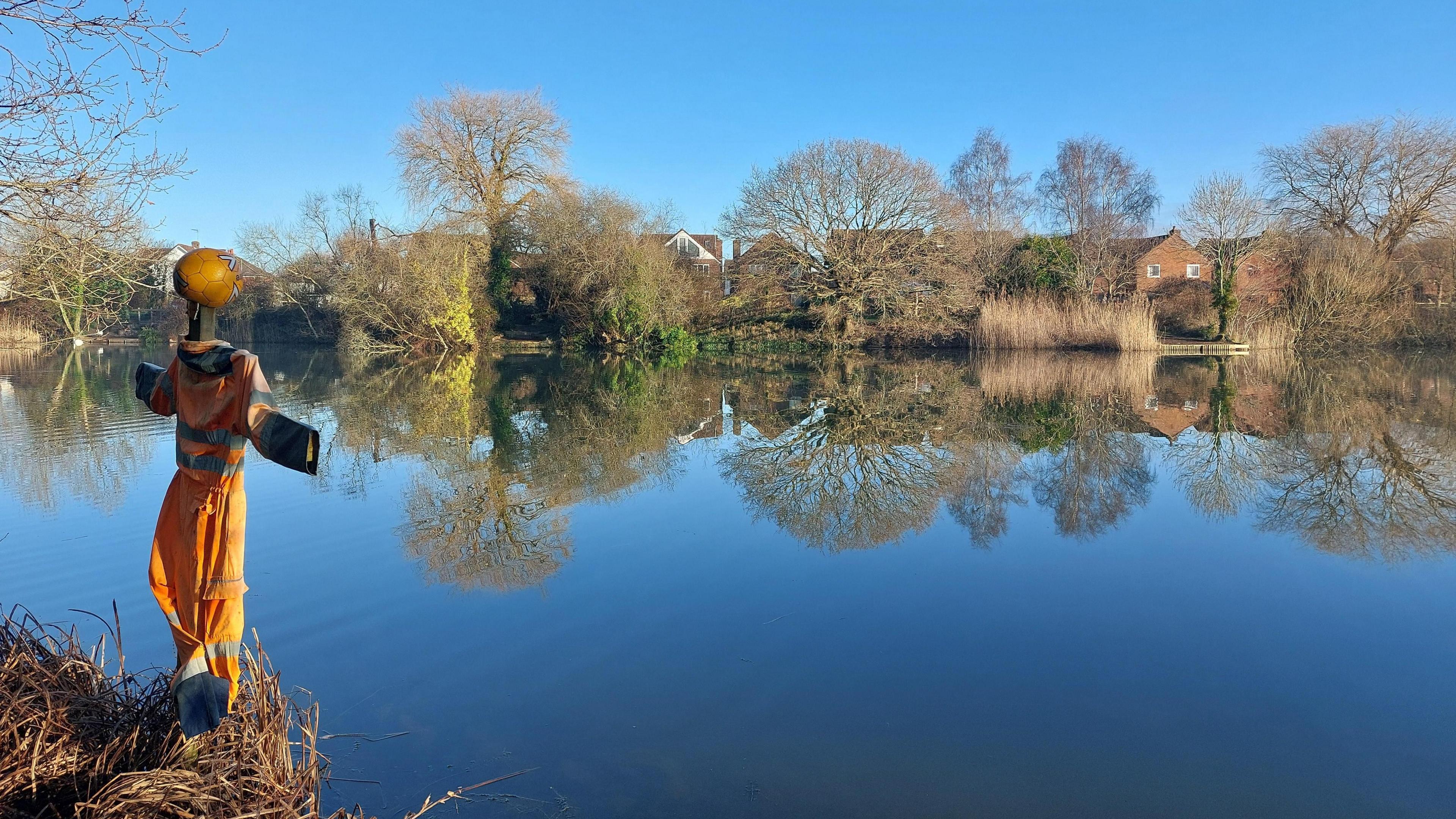 A scarecrow type character made of a wooden cross with an orange jump suit and helmet is on the bank of a body of water. It is a sunny winters day. On the far bank you can see brick homes through trees. 