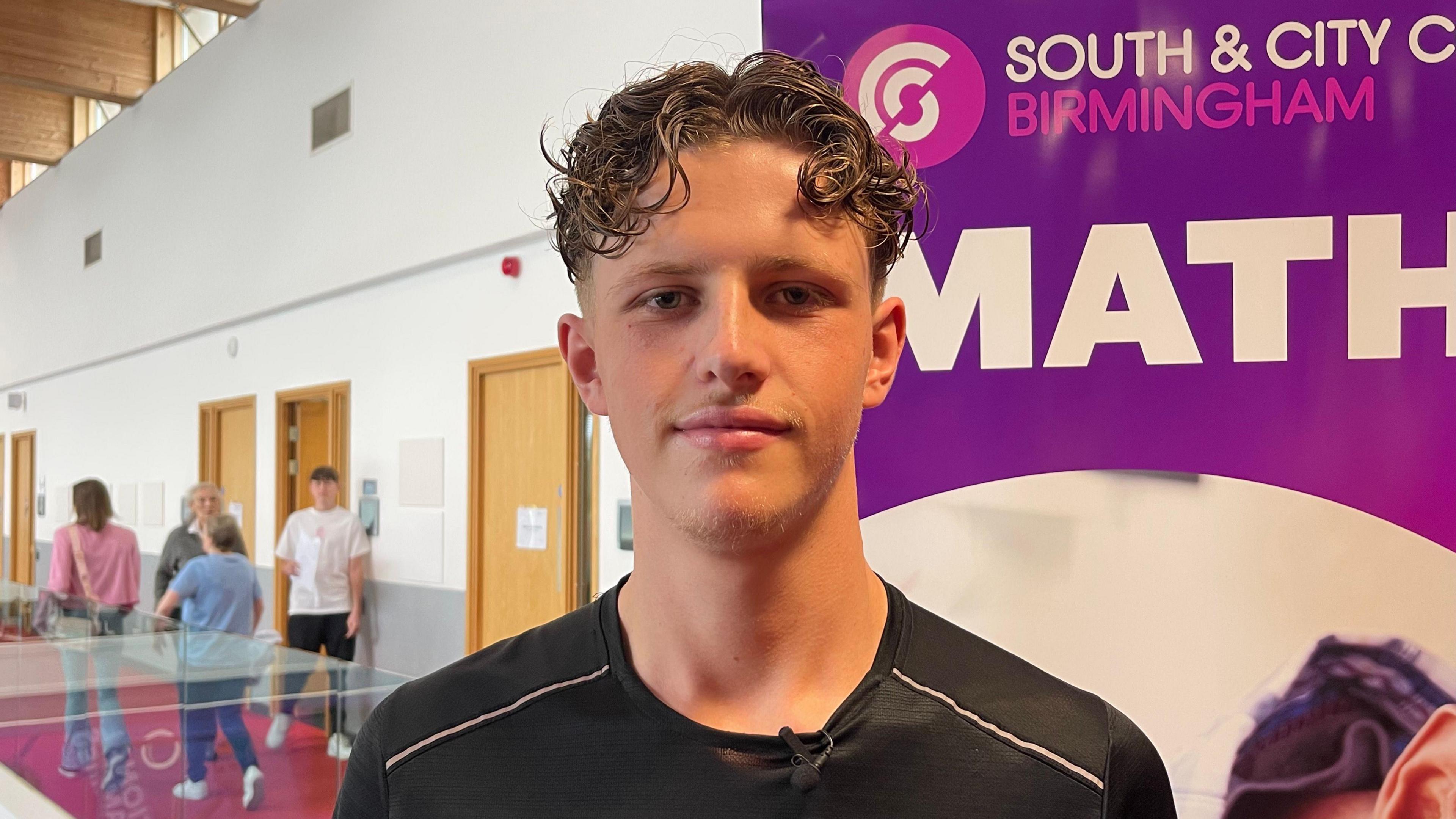 James Bonning, a young man with curly brown hair, stands in a school room in front of a sign saying "South & City College Birmingham". He's wearing a black sports T-shirt and is smiling at the camera. 