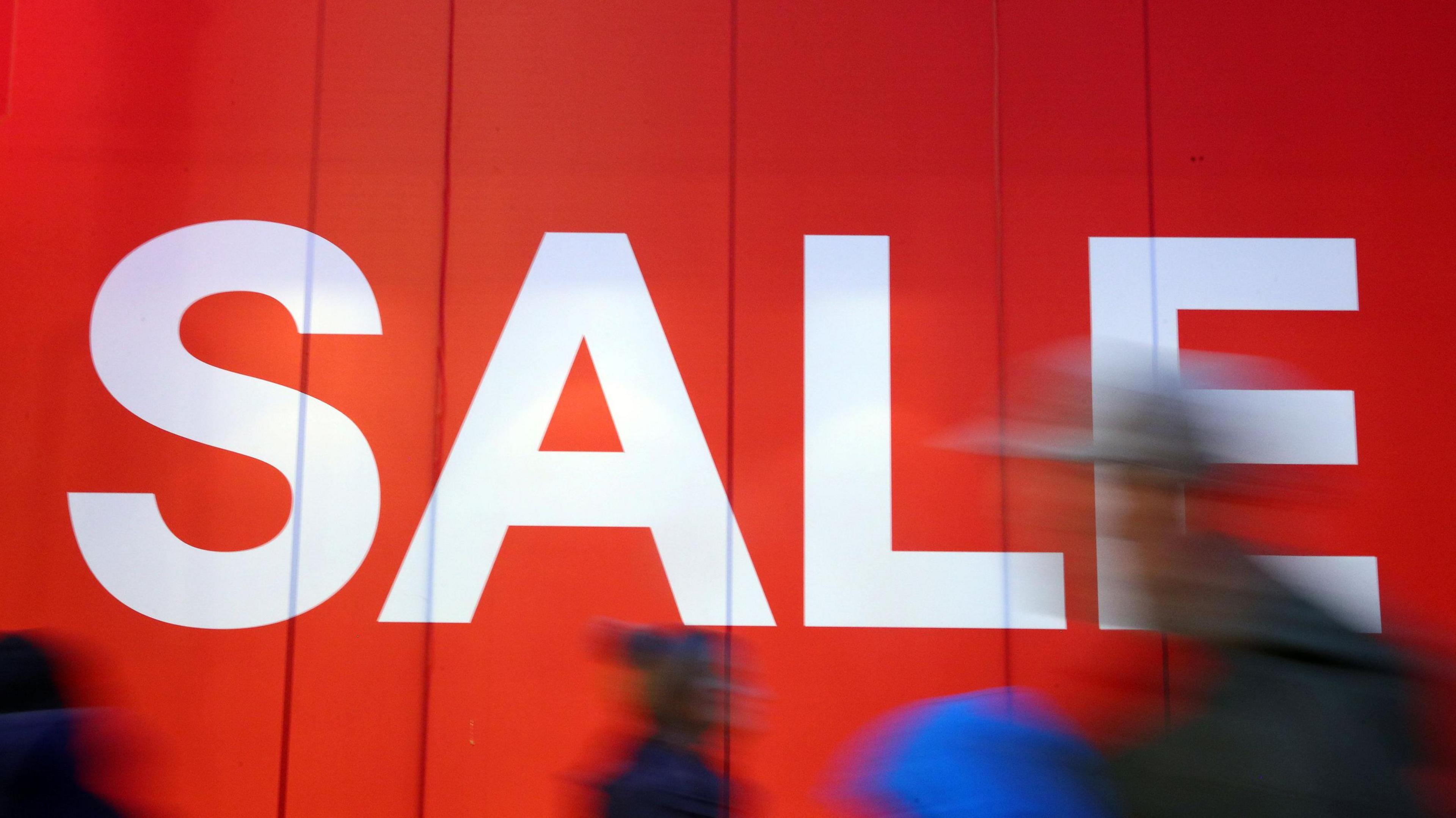 Shoppers walking in front of a large red sign that says "sale" in white font.