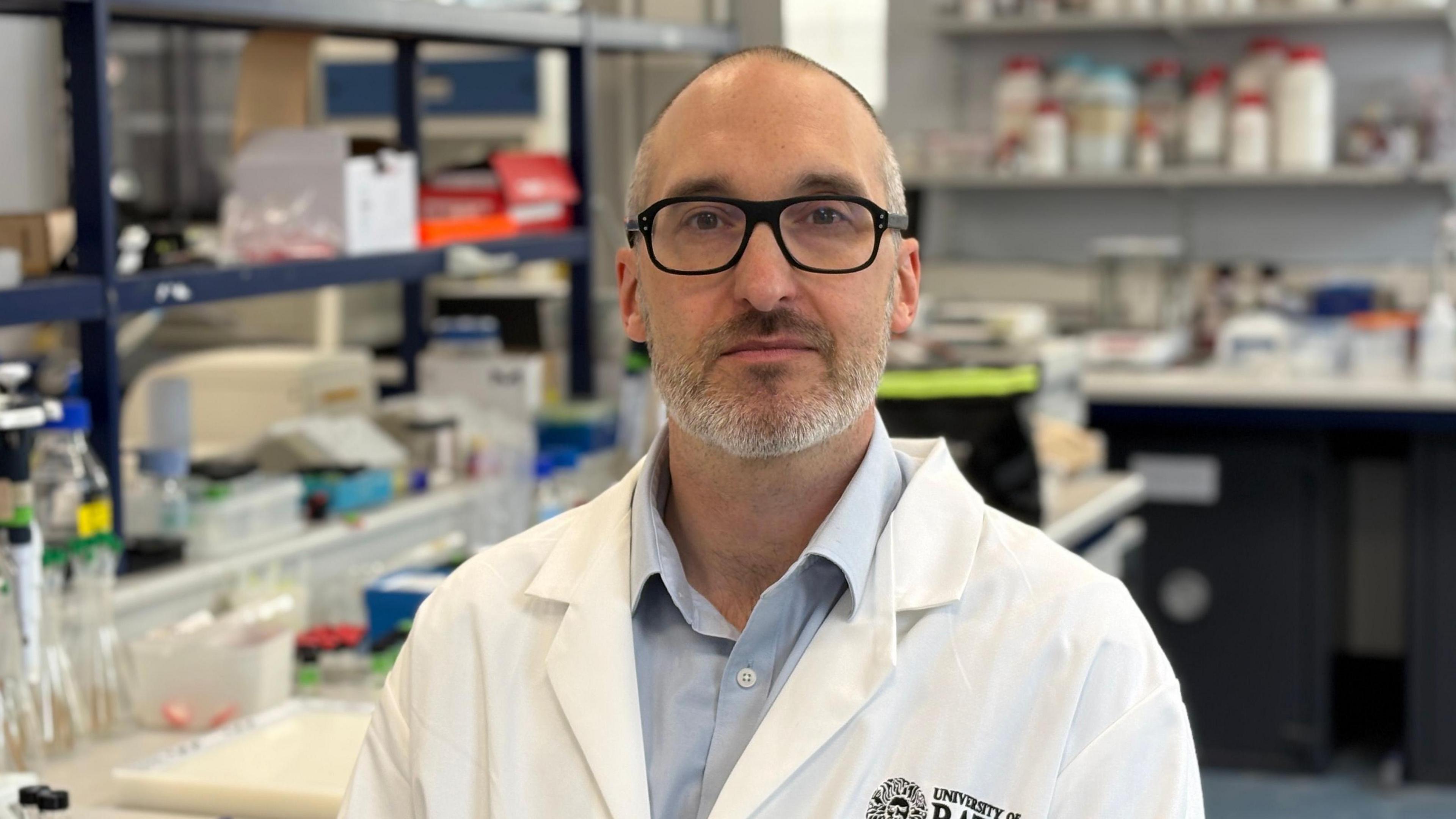 Professor Chris Pudney is in a lab with a white lab coat on over a blue shirt. He is wearing black-rimmed glasses and he has grey receding hair and grey stubble.