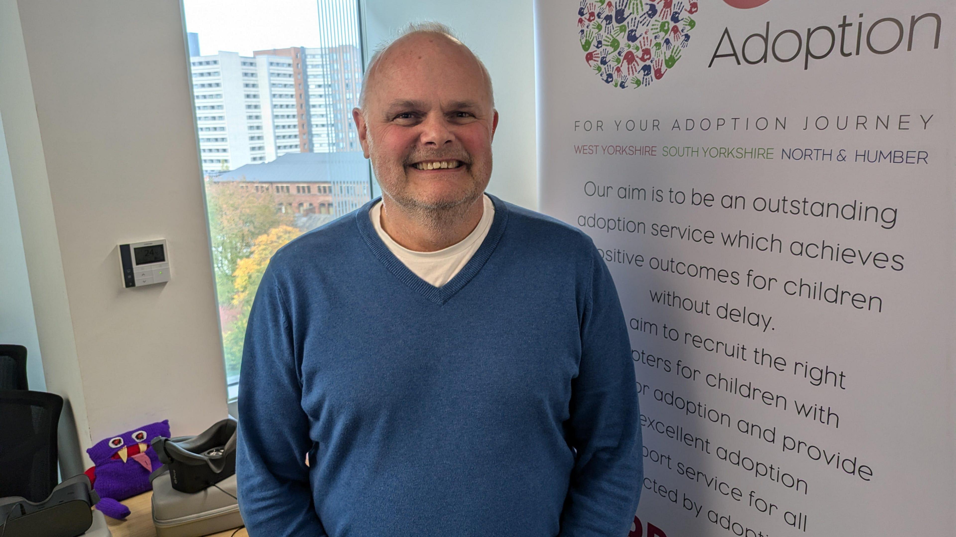 A man in his fifties wearing a blue V-neck pullover. He is stood next to an advertising board for One Adoption, which has large writing on explaining what the agency is. A window is behind him. 