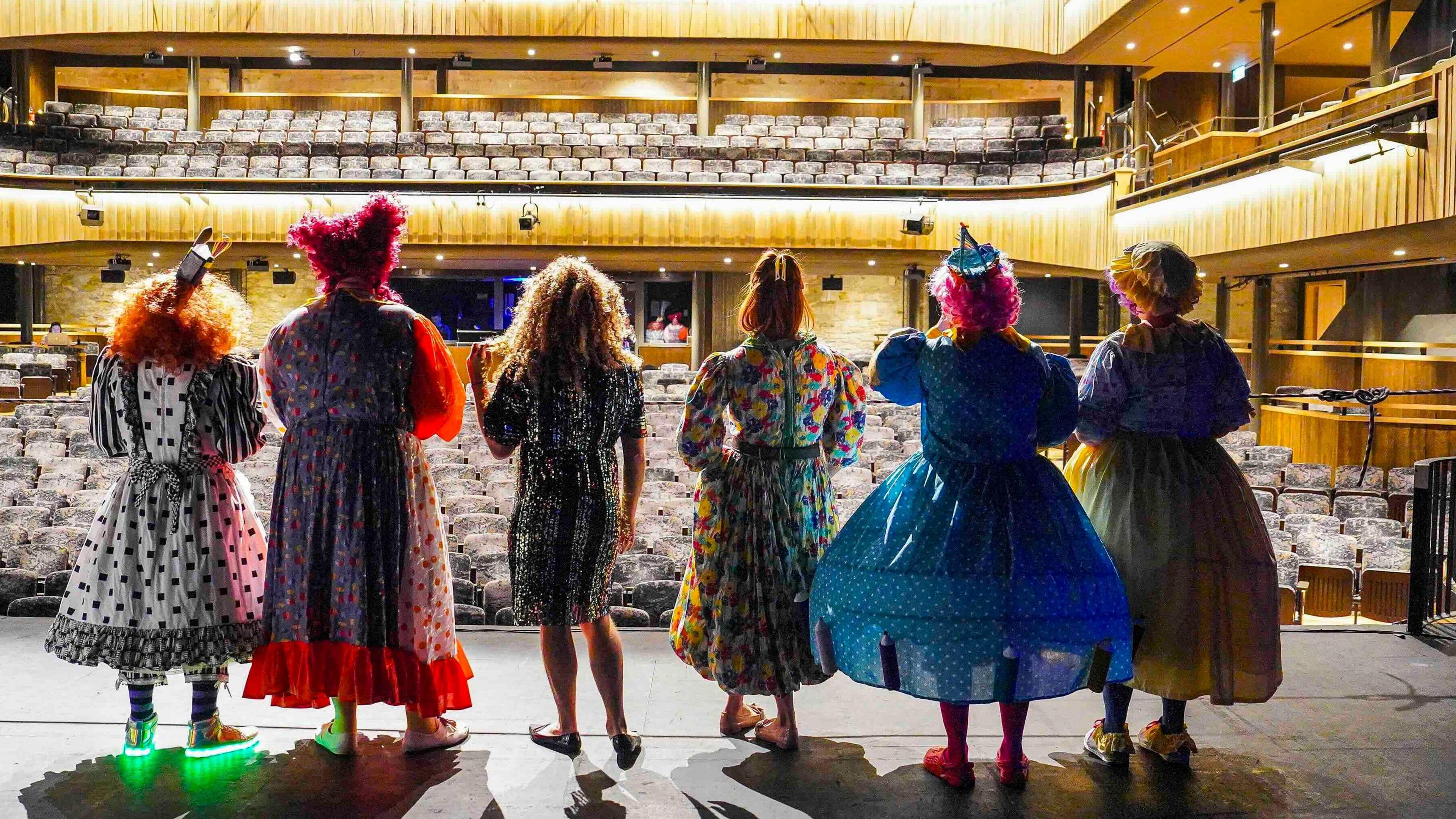 A view of six pantomime dames from behind with a theatre auditorium in front of them.