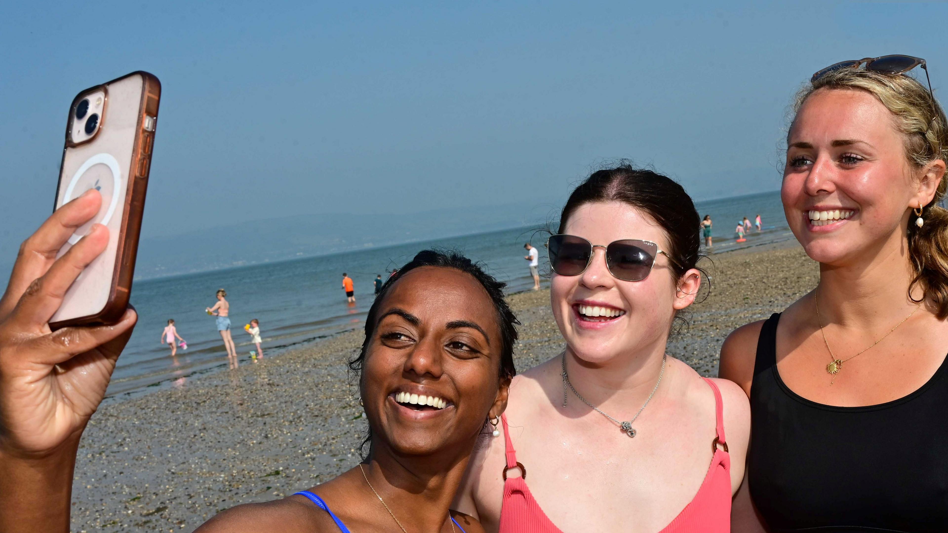 Three girls on a beach taking a selfie. They are all in swimsuits. Two have dark hair and one has blond hair. They are all smiling for the camera.