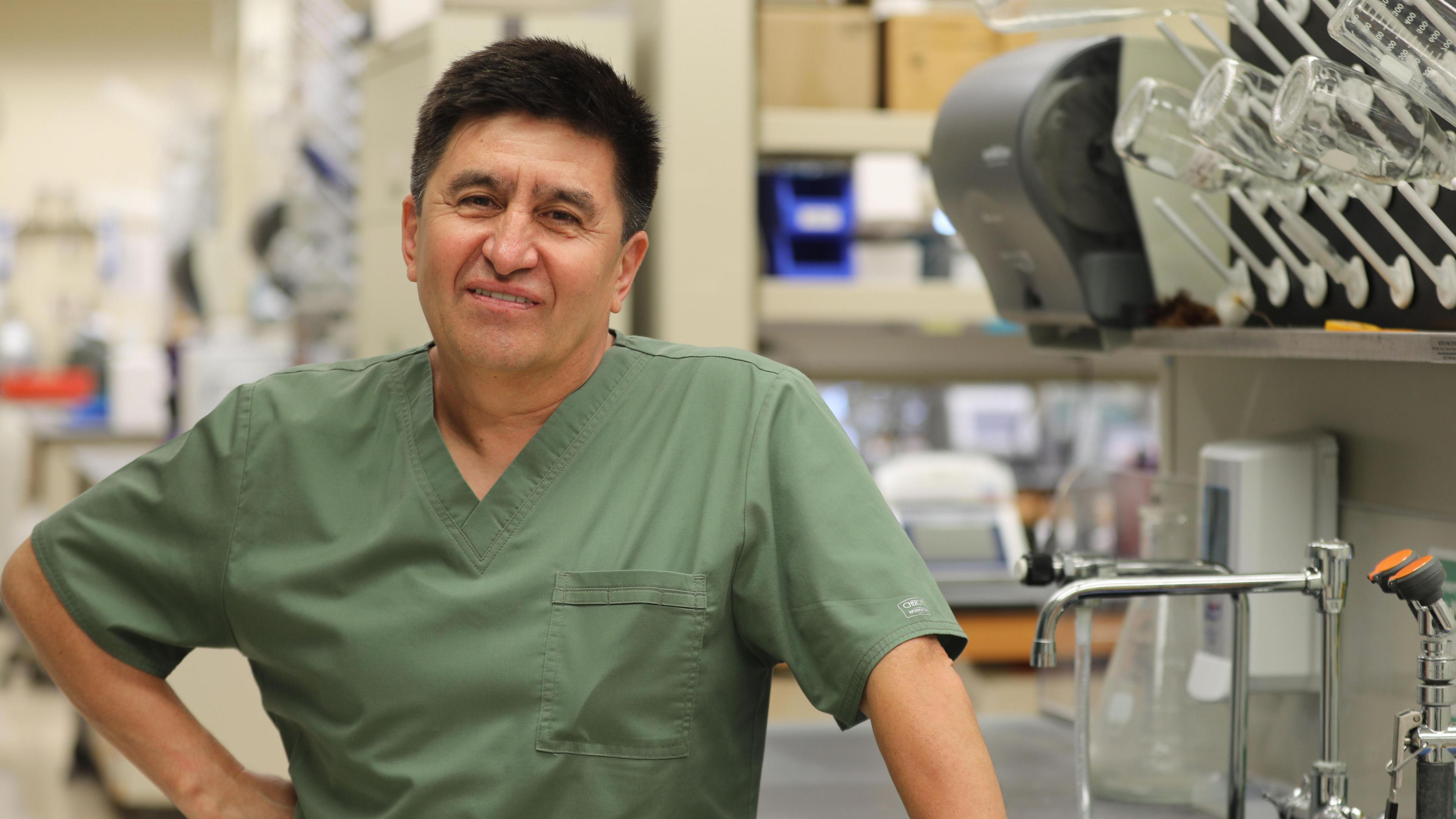 Man in olive-green scrubs in a laboratory with taps and scientific equipment in the background, smiling at the camera.