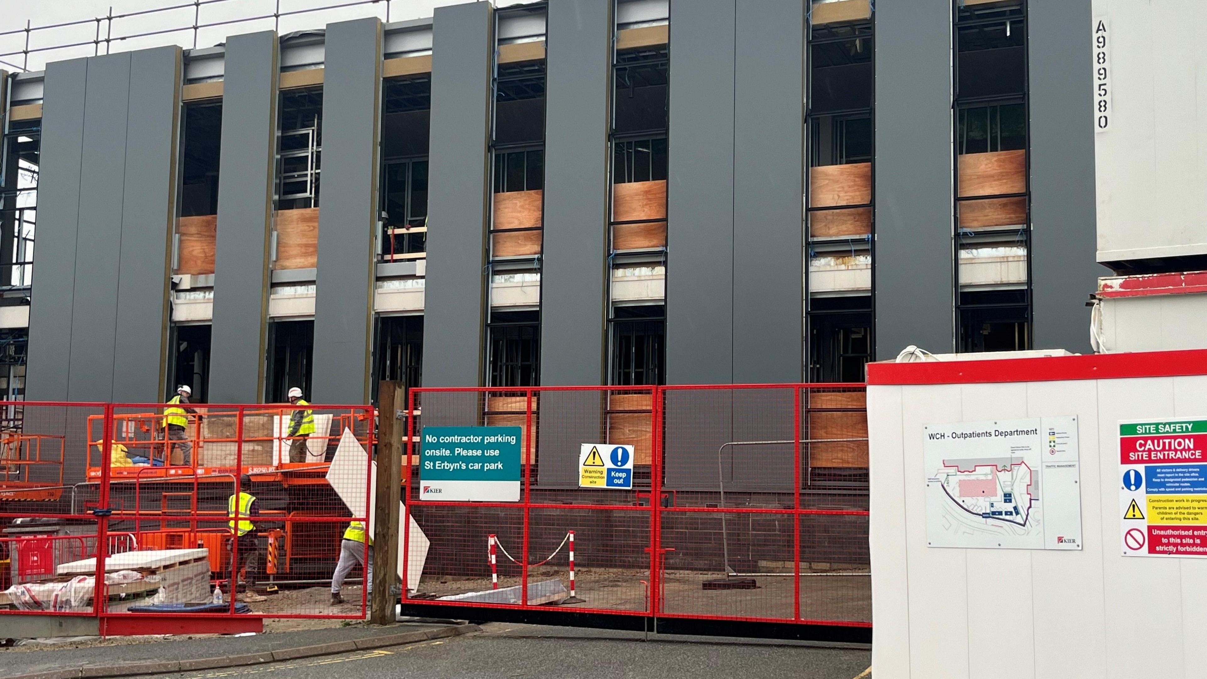 Four men wearing high-viz jackets are working on the new building under construction. It has two storeys and the walls are incomplete, with gaps showing. There is a red fence around the building site and a portacabin with safety warnings and a site map on it in the foreground.