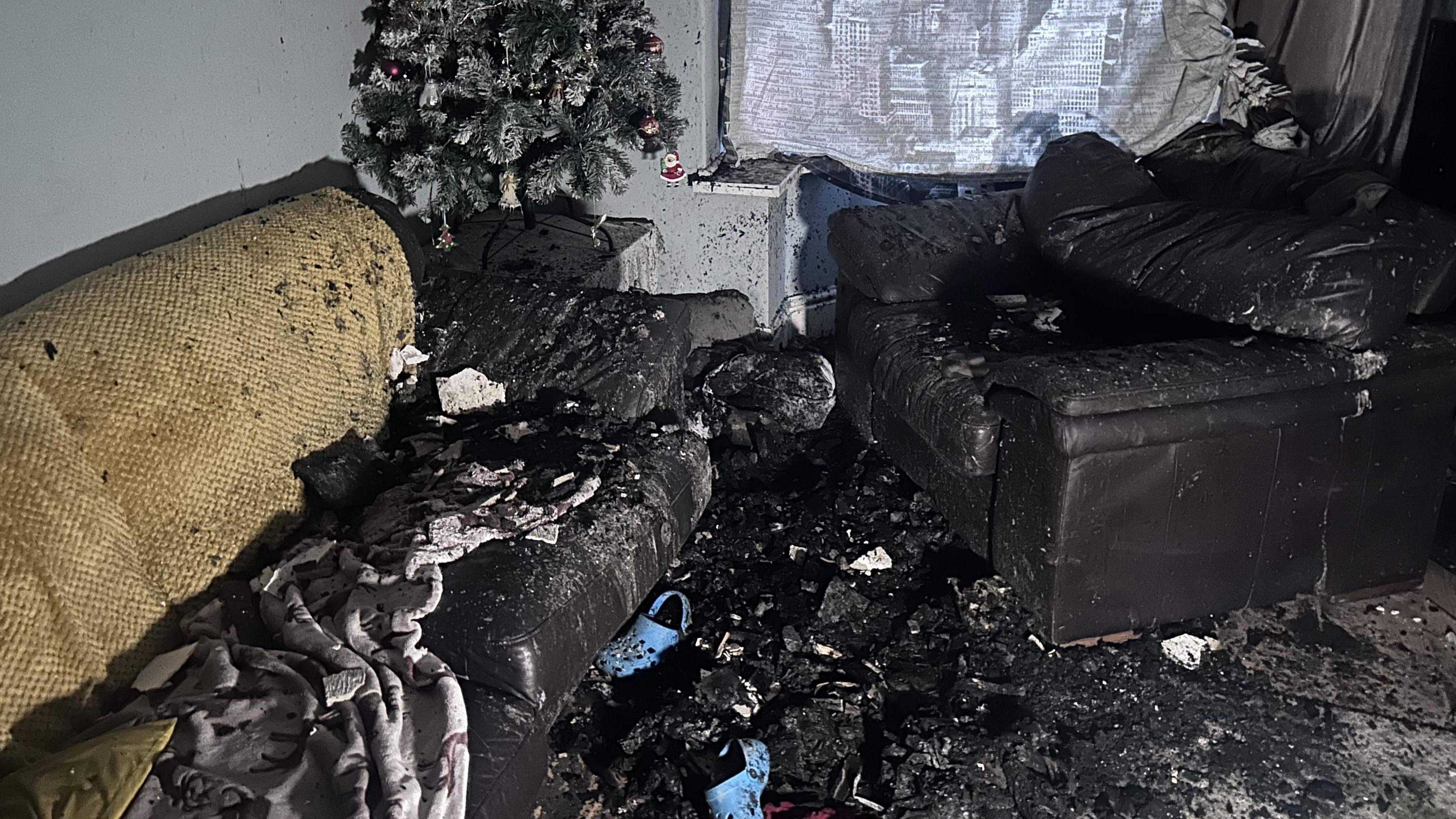 A living room with severe fire damage, lots of dust and ash and blacked out furniture. A fire-damaged Christmas tree is in the corner.