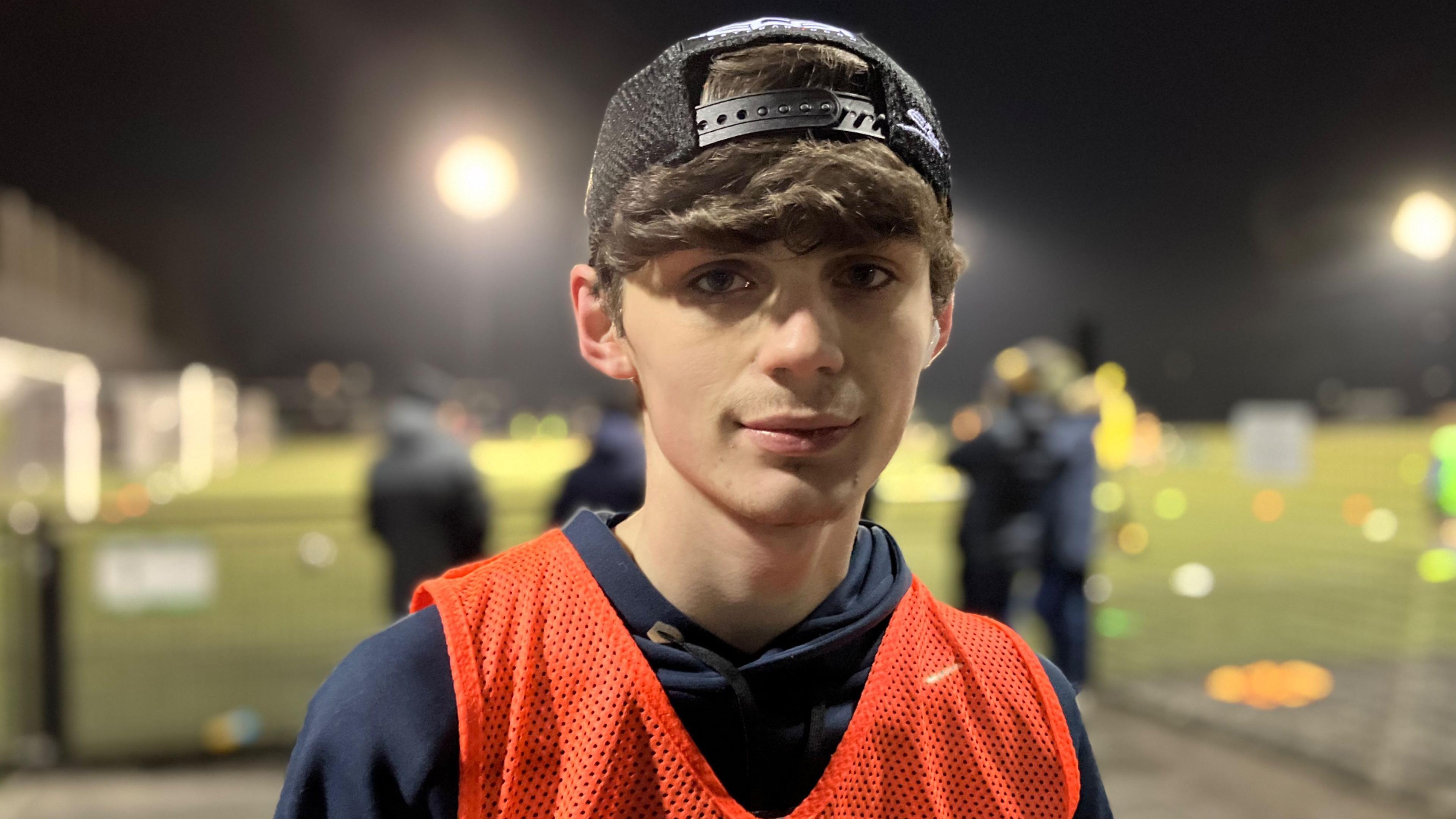 Luke looks into the camera in the centre of the image, with blurred astro football pitches lit by floodlights in the background. He has a backwards cap on with hair down over his forehead,  and he is wearing an orange bib over his hoodie.