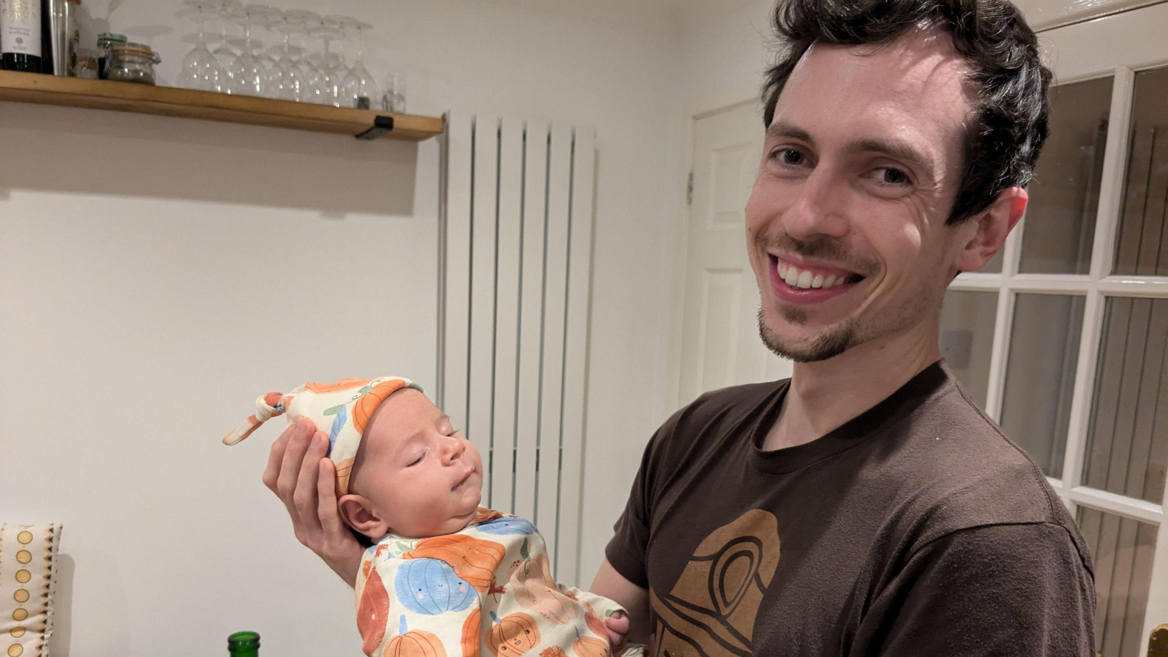 A man in a brown t-shirt smiles at the camera while holding his baby son