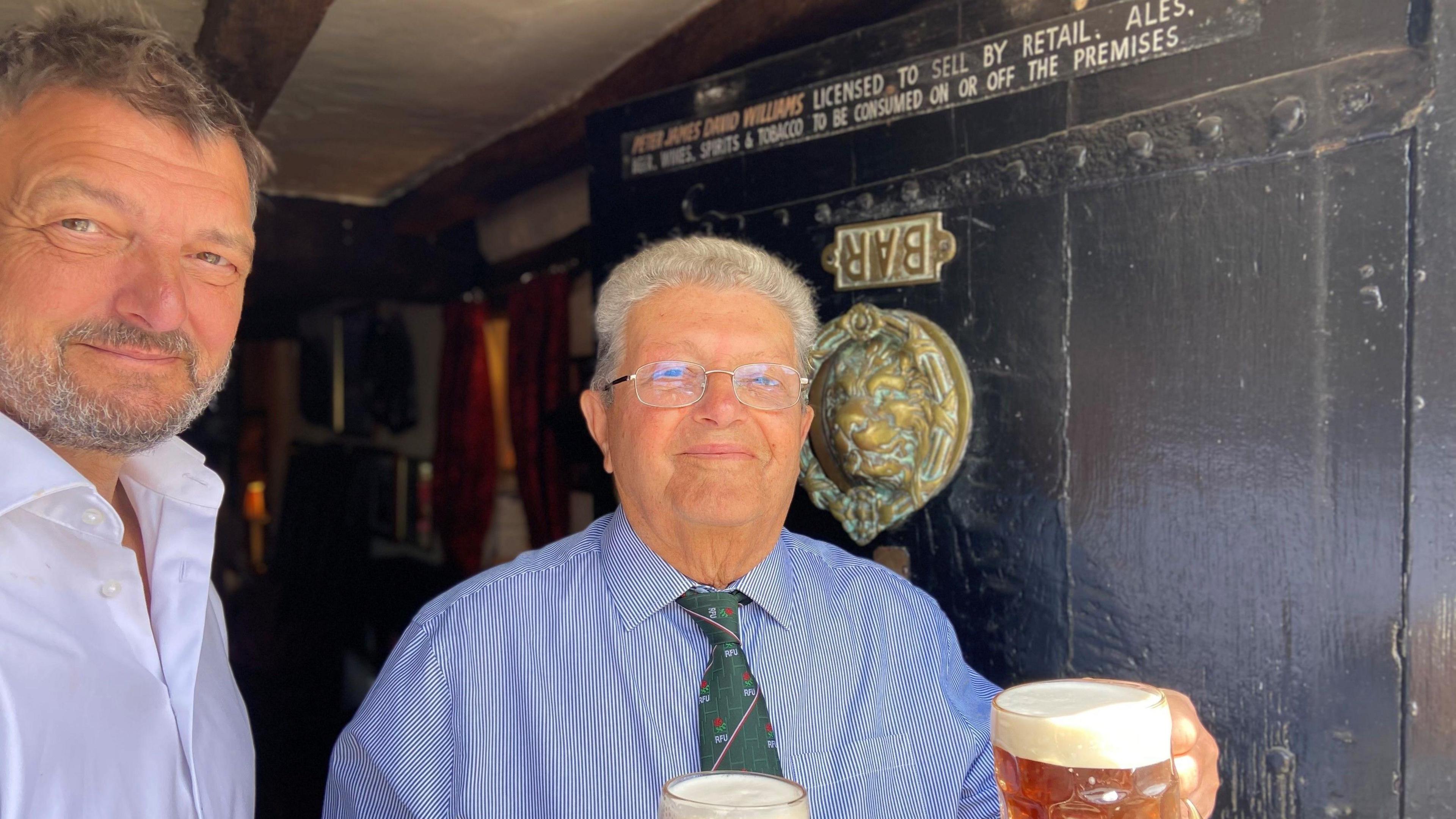 A man with grey hair and glasses is holding a cup of beer in front of a door. The door has an upside-down sign that reads "bar". There is a large brass lion face on the door. Next to the man is another man with dark hair and grey facial hair