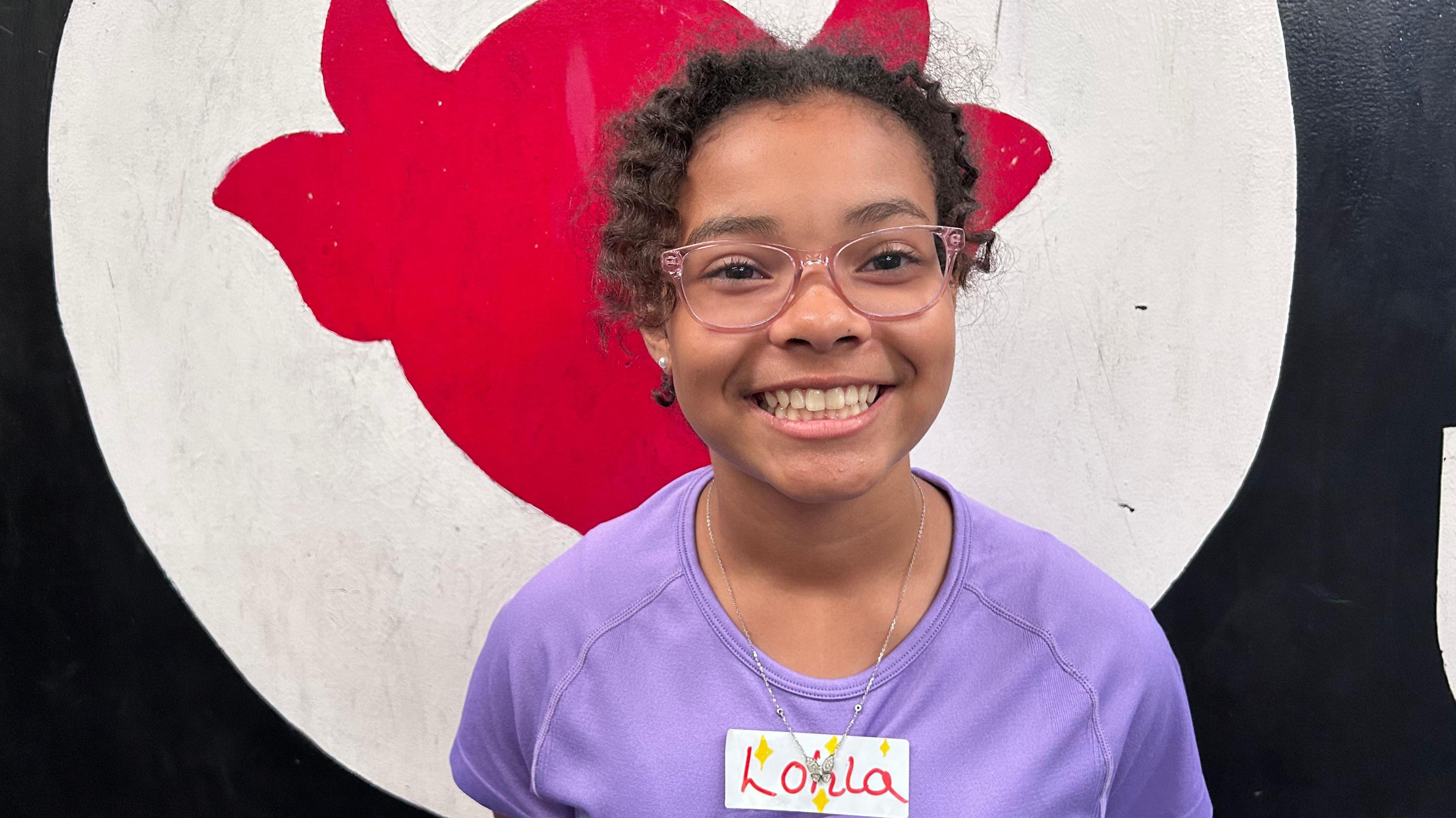 Lolah is wearing a name badge, lilac t shirt and pink framed glasses, she is smiling toward the camera