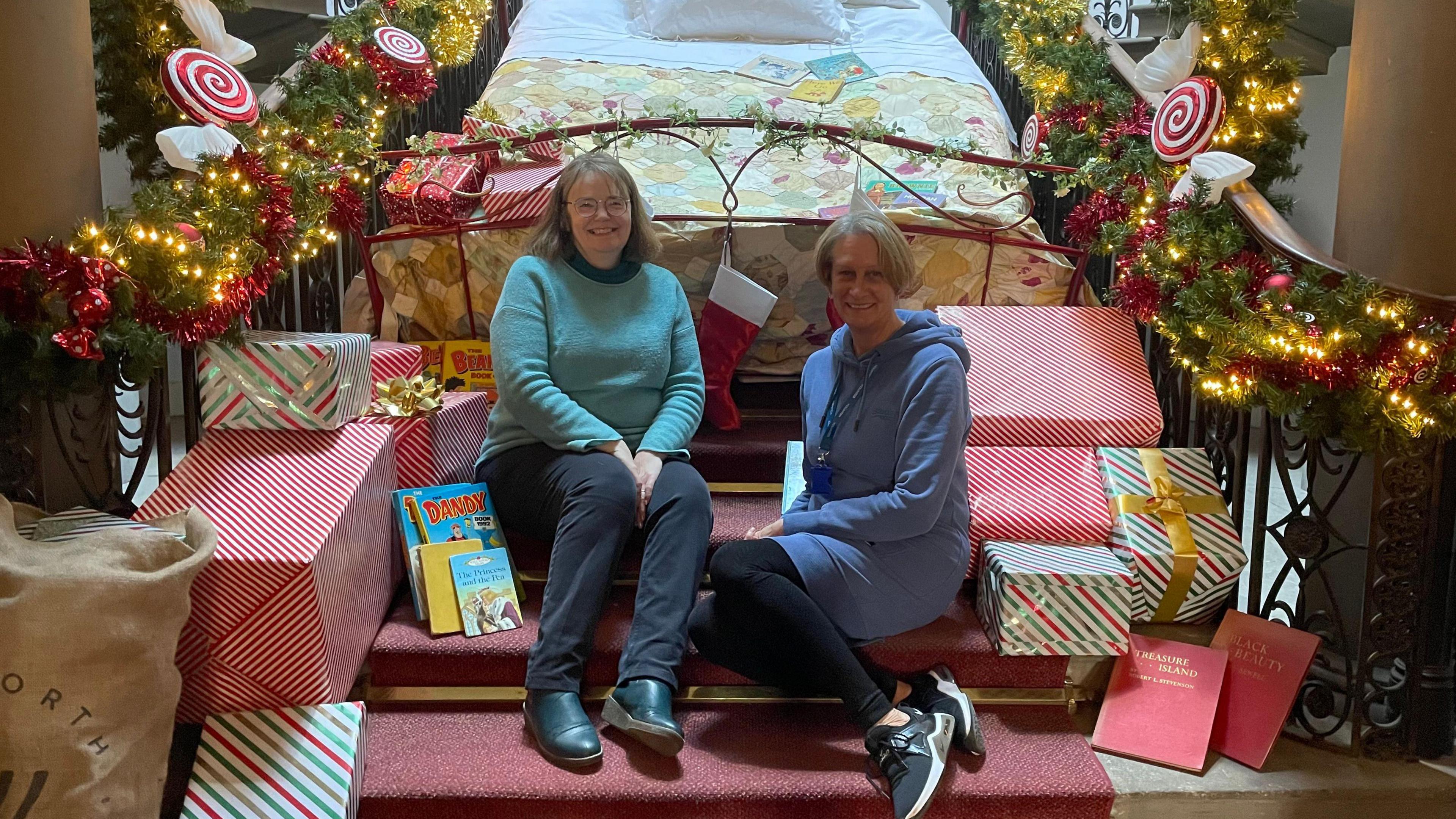 Victoria and Julie are smiling whilst sitting next to each other on a decorated staircase. All around them are wrapped presents, but behind them, a bed is balanced on the stairs with stockings hanging from it. The bannisters are wrapped with garland, tinsel, and lights.