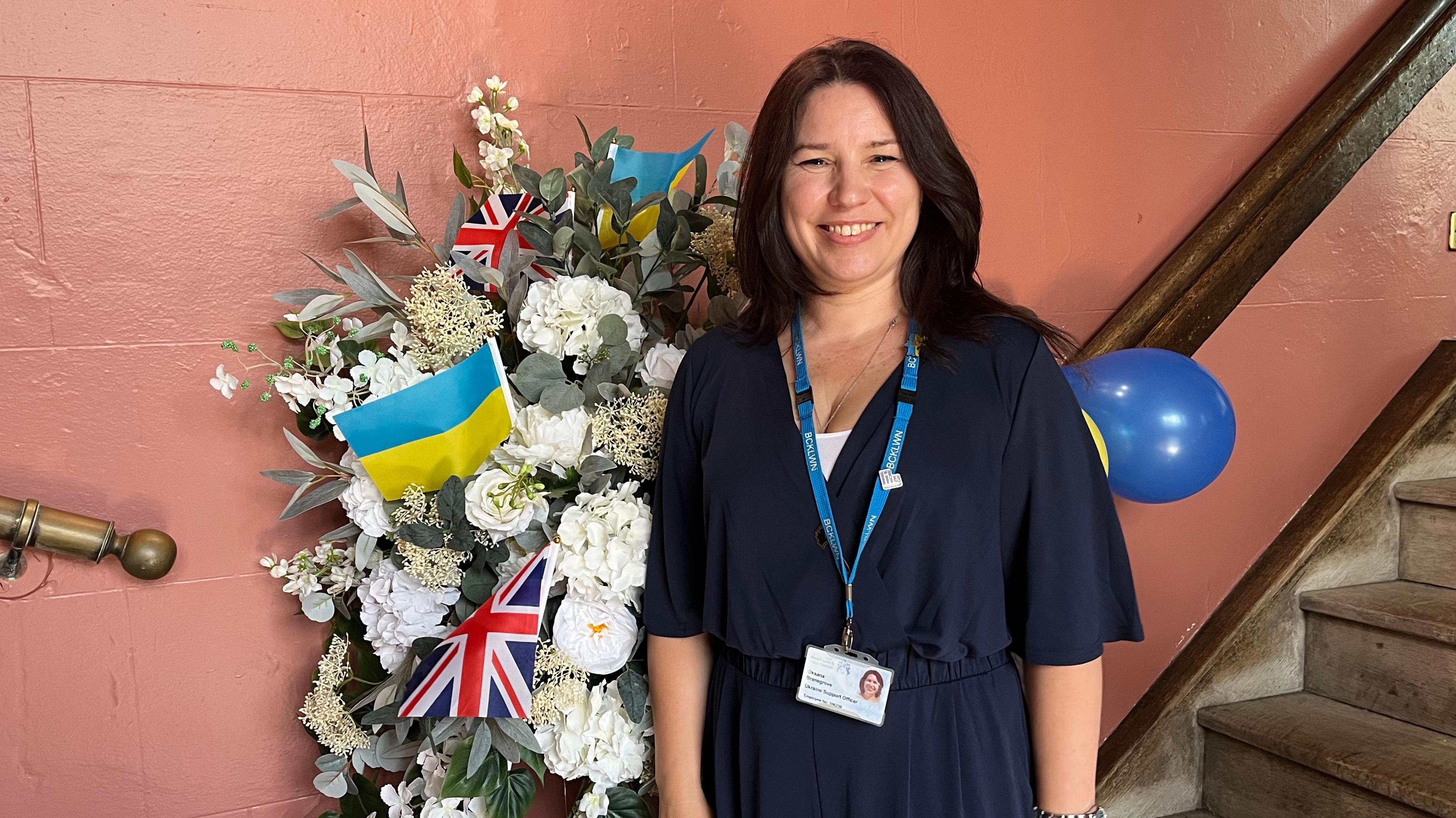 Oksana Bransgrove, Ukraine Support Officer at West Norfolk Borough Council, is wearing a navy jump suit and has her hair in a brown bob. She stands in front of a large bunch of white flowers decorated with British and Ukrainian flags and blue and yellow balloons. 