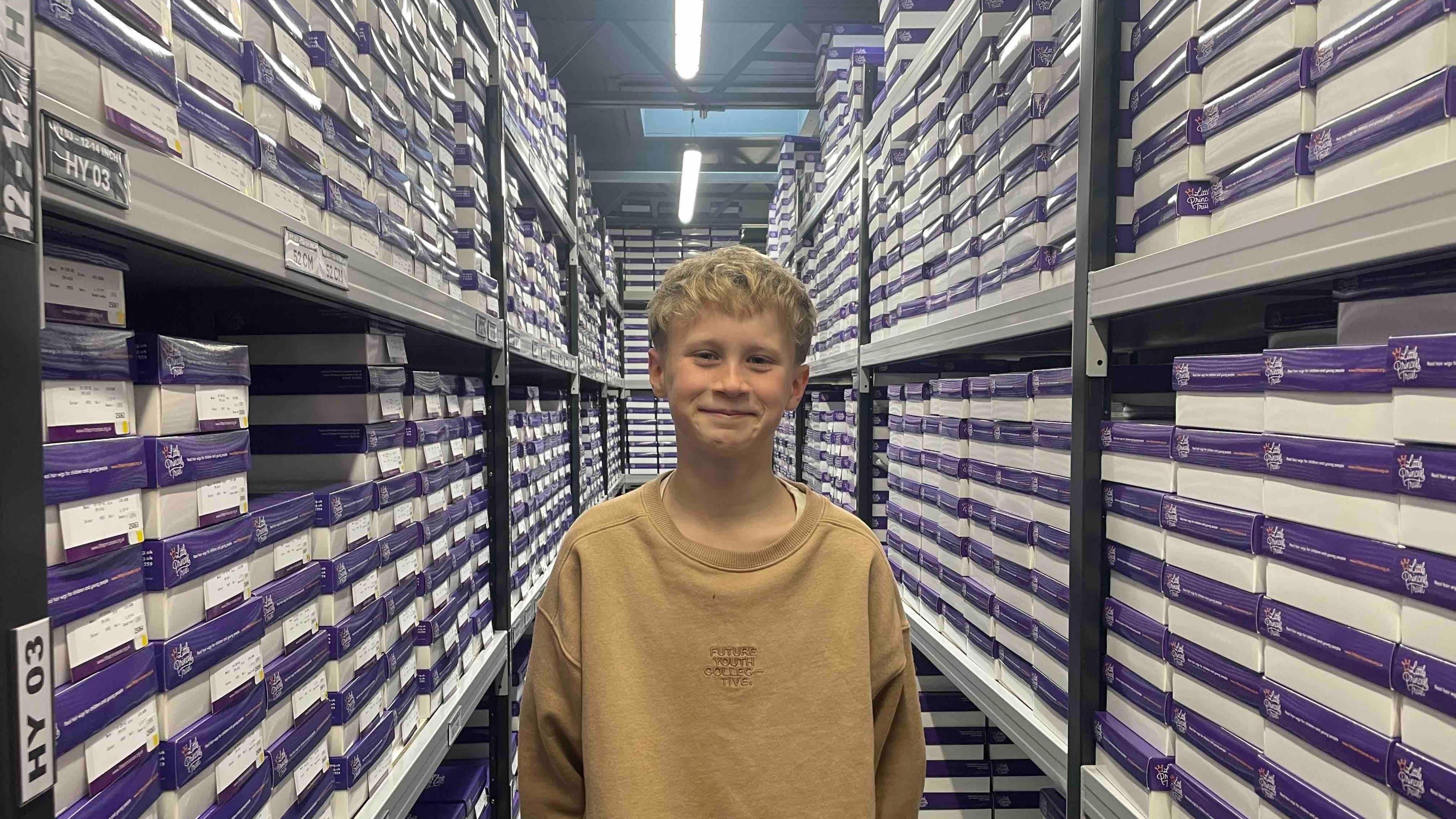 A blond boy wearing a brown sweatshirt is stood in a warehouse setting with boxes on either side of him.