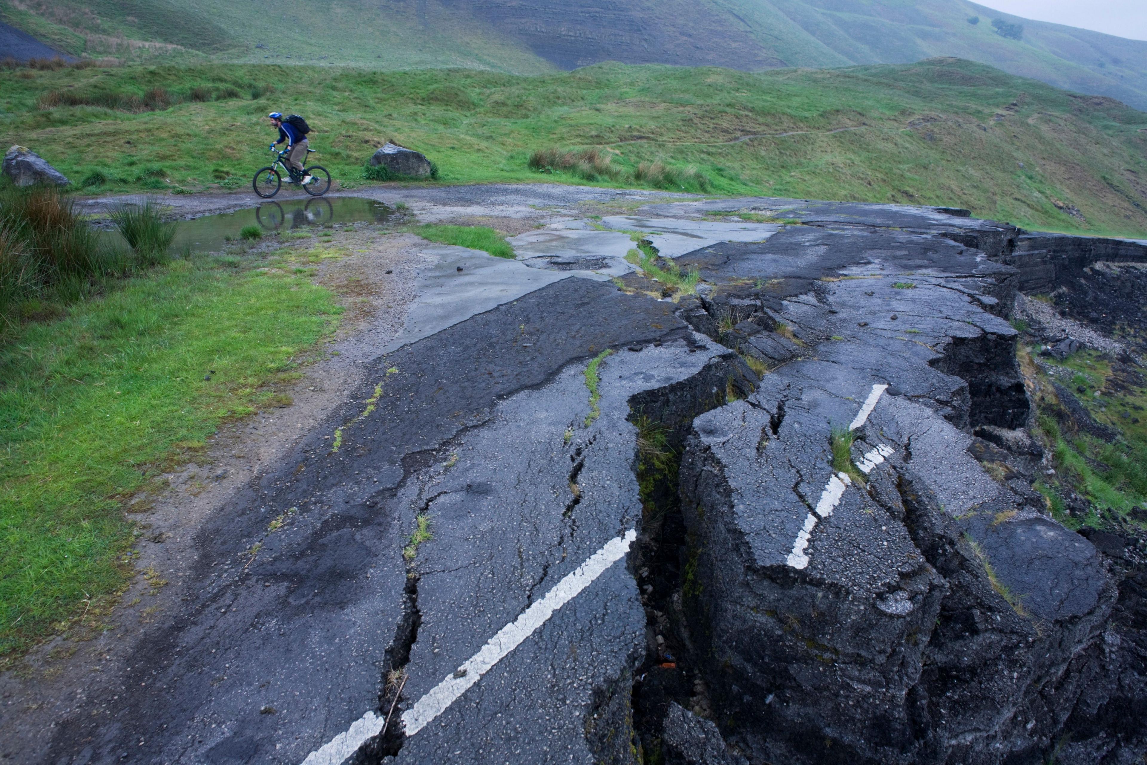 Broken asphalt, with large cracks in it, and painted white lines, indicating where a road used to run near Mam Tor.