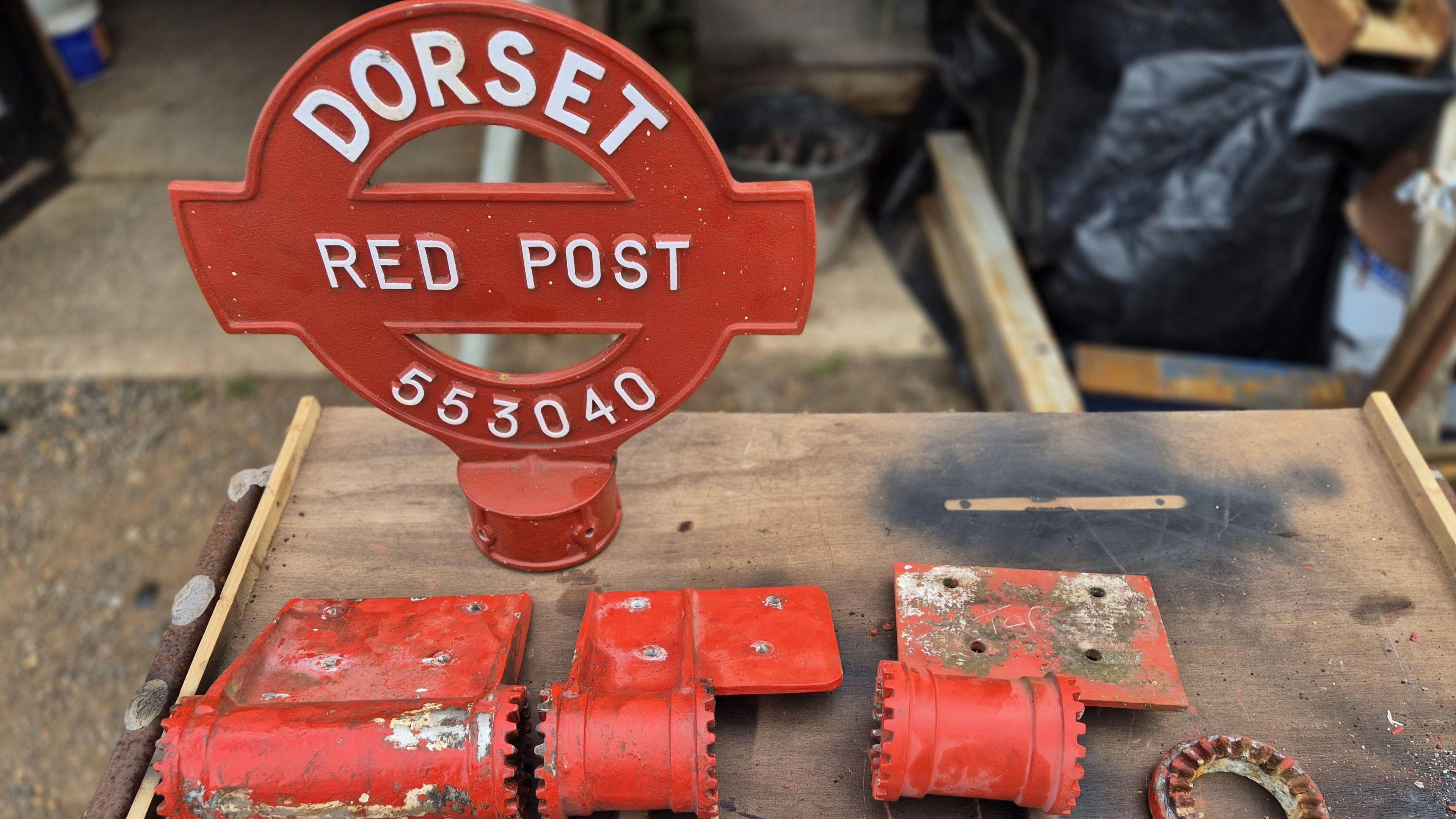 A red fingerpost is shown prior to its restoration, broken down into its parts and laid out on a workbench. The post's head reads DORSET RED POST with a grid reference number underneath ,and several cast iron bolts and panels, also in red, laid out next to it. 