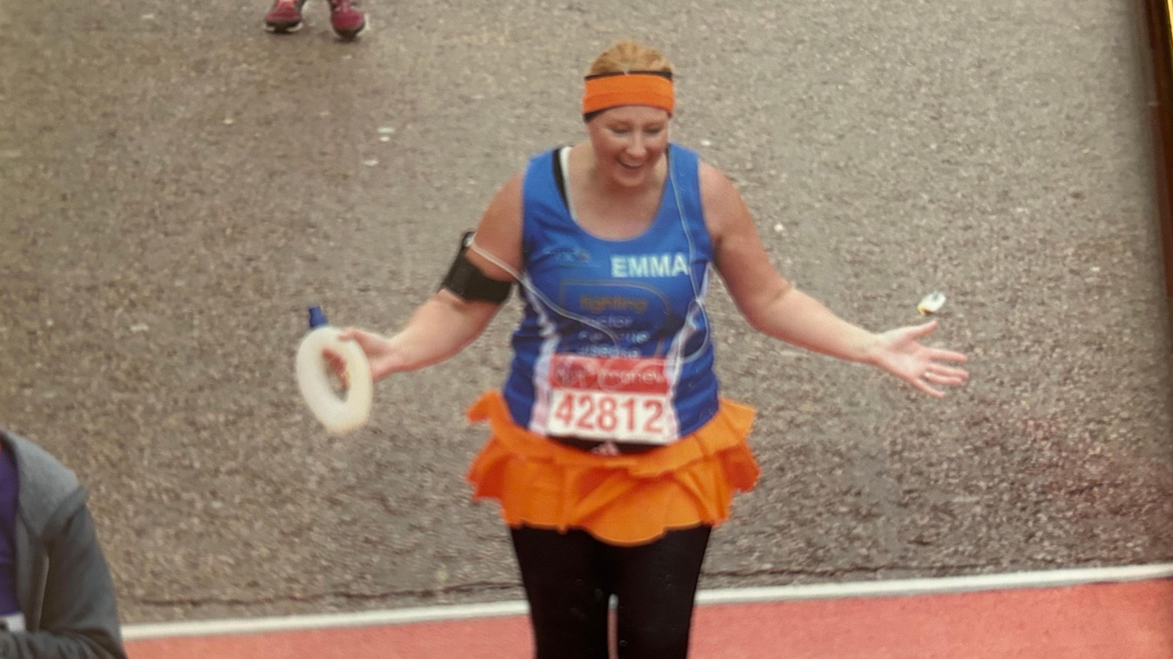 A woman dressed in a blue running top, black leggings and orange headband crosses a finish line holding a circular water bottle