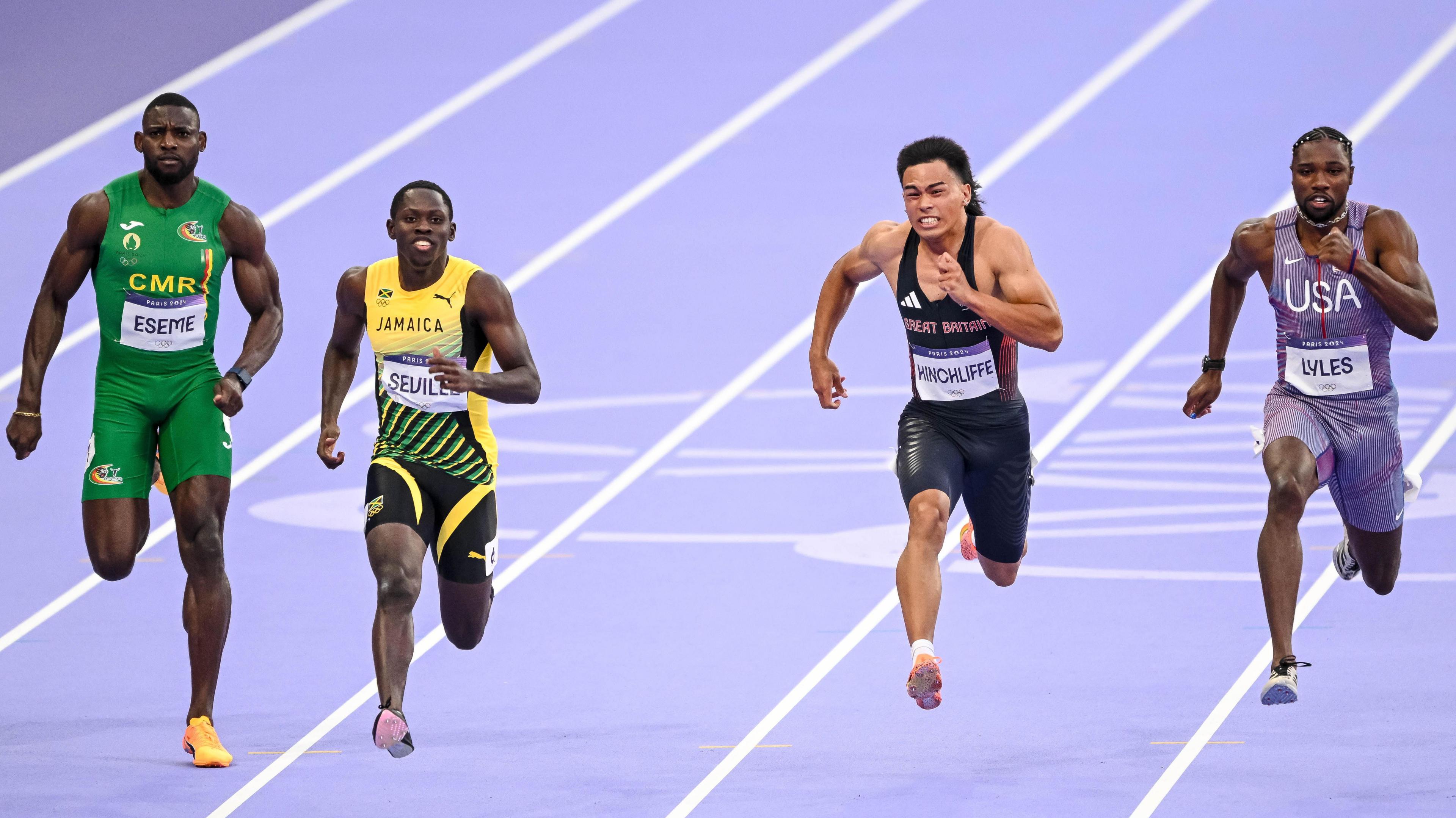 An action shot from a men's 100m semi-final at the Paris 2024 Olympics sees four men straining while racing on a purple track, with three of the runners with both feet off the ground. From left to right, the runners are: Emmanuel Eseme of Cameroon, Oblique Seville of Jamaica, Louie Hinchliffe of Great Britain and Noah Lyles of USA
