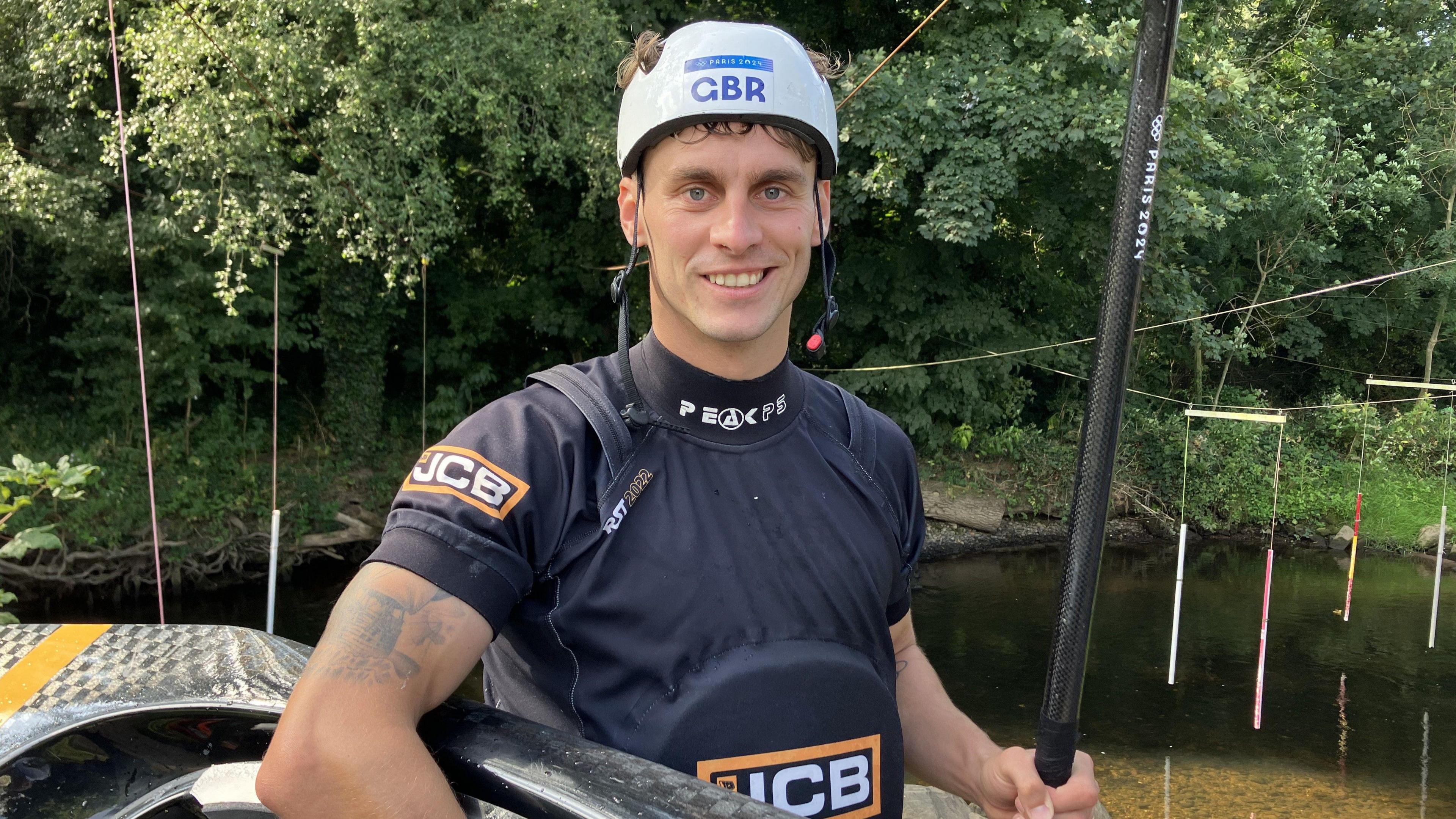 Adam Burgess is wearing a black t-shirt and a white helmet. He is holding a canoe paddle and a canoe. In the background is a river.