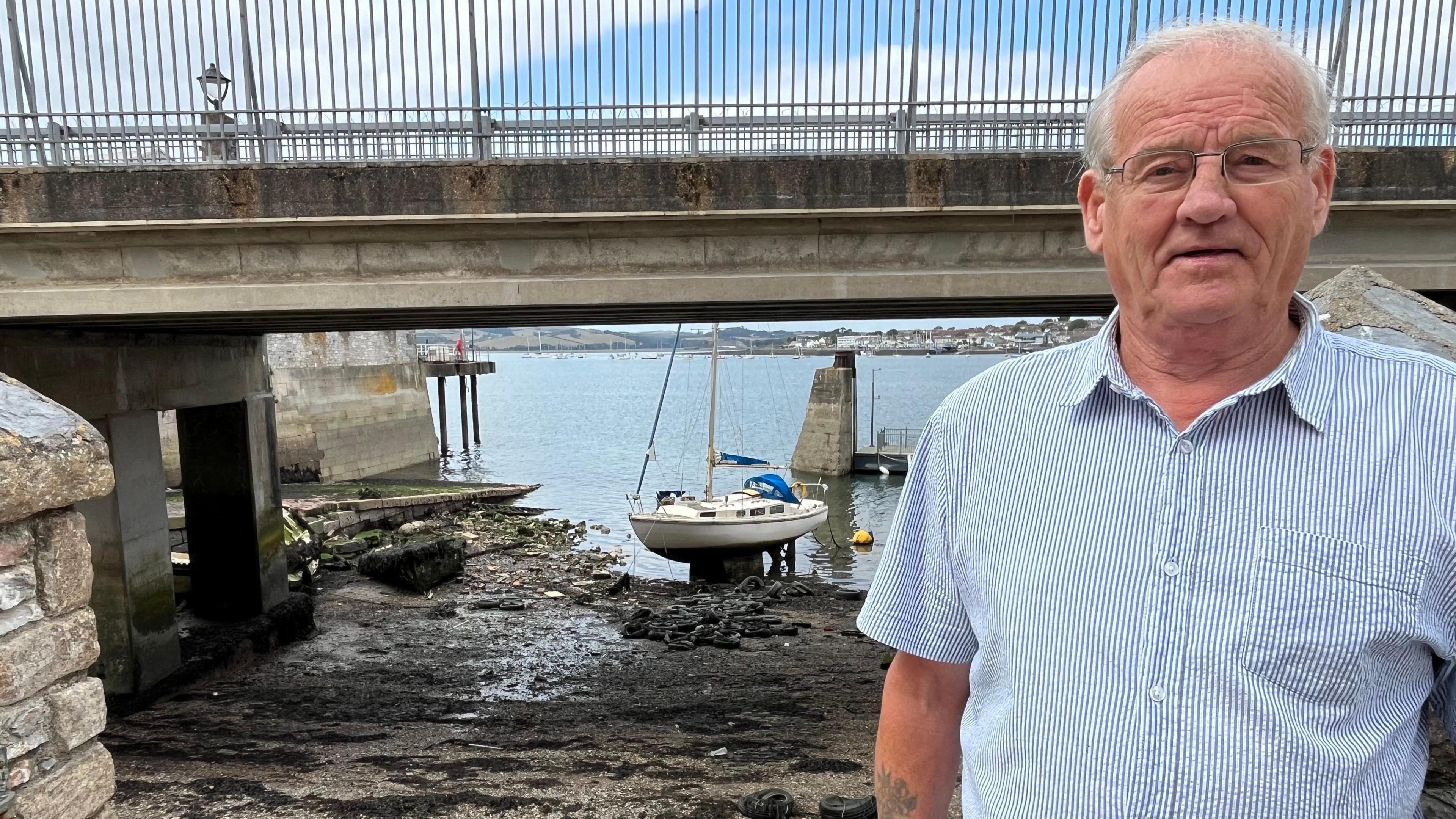 Phil Head, wearing a blue and white striped shirt and glasses. He is stood in the right of the frame. Behind him is a beach and a boat. The water is blue. The sky is blue and filled with clouds.