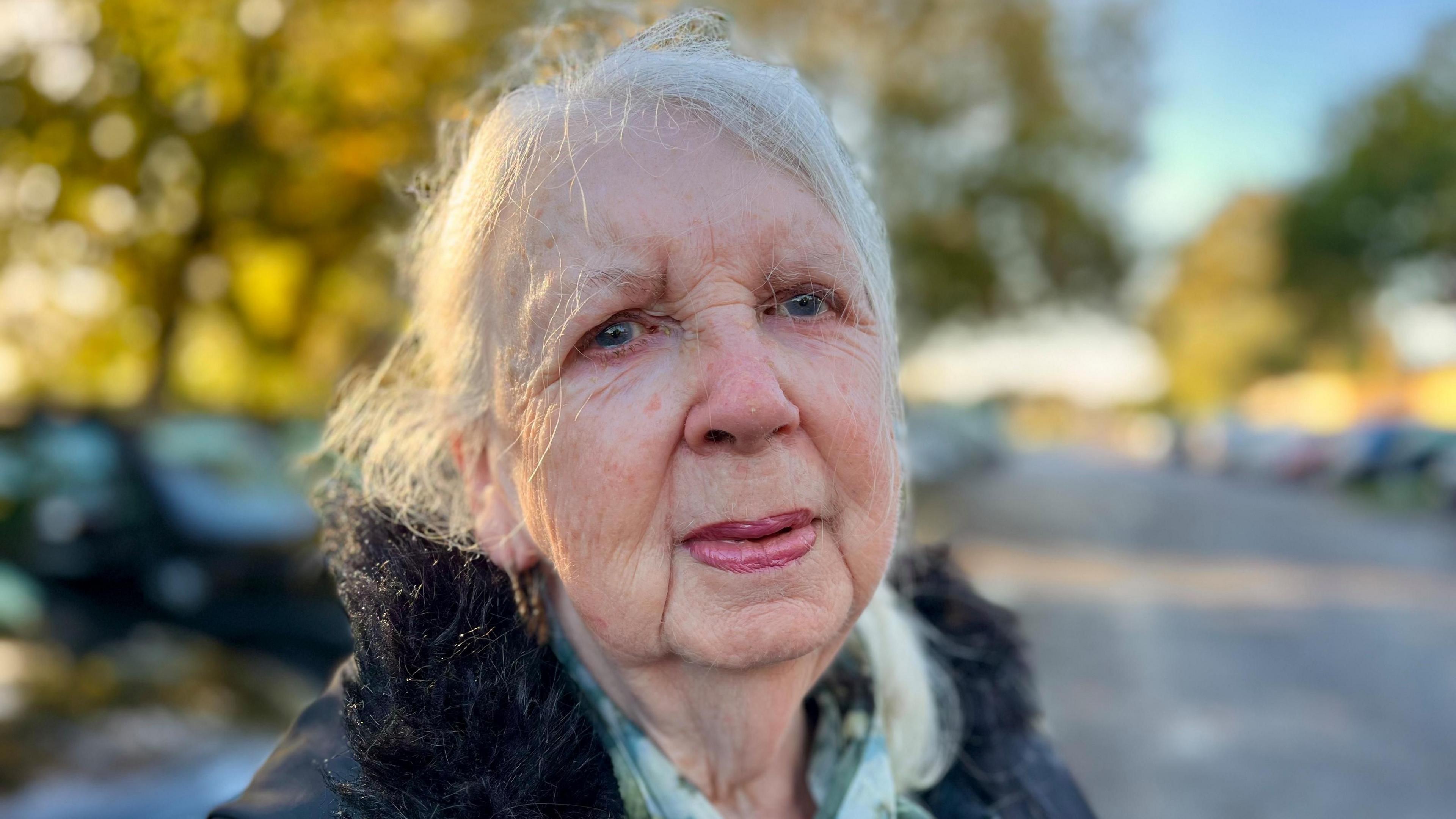 Brenda has long white hair pulled back from her face. She is wearing a black jacket with a furry collar and a green checked shirt underneath.
She is looking at the camera with trees and parked cars behind her.