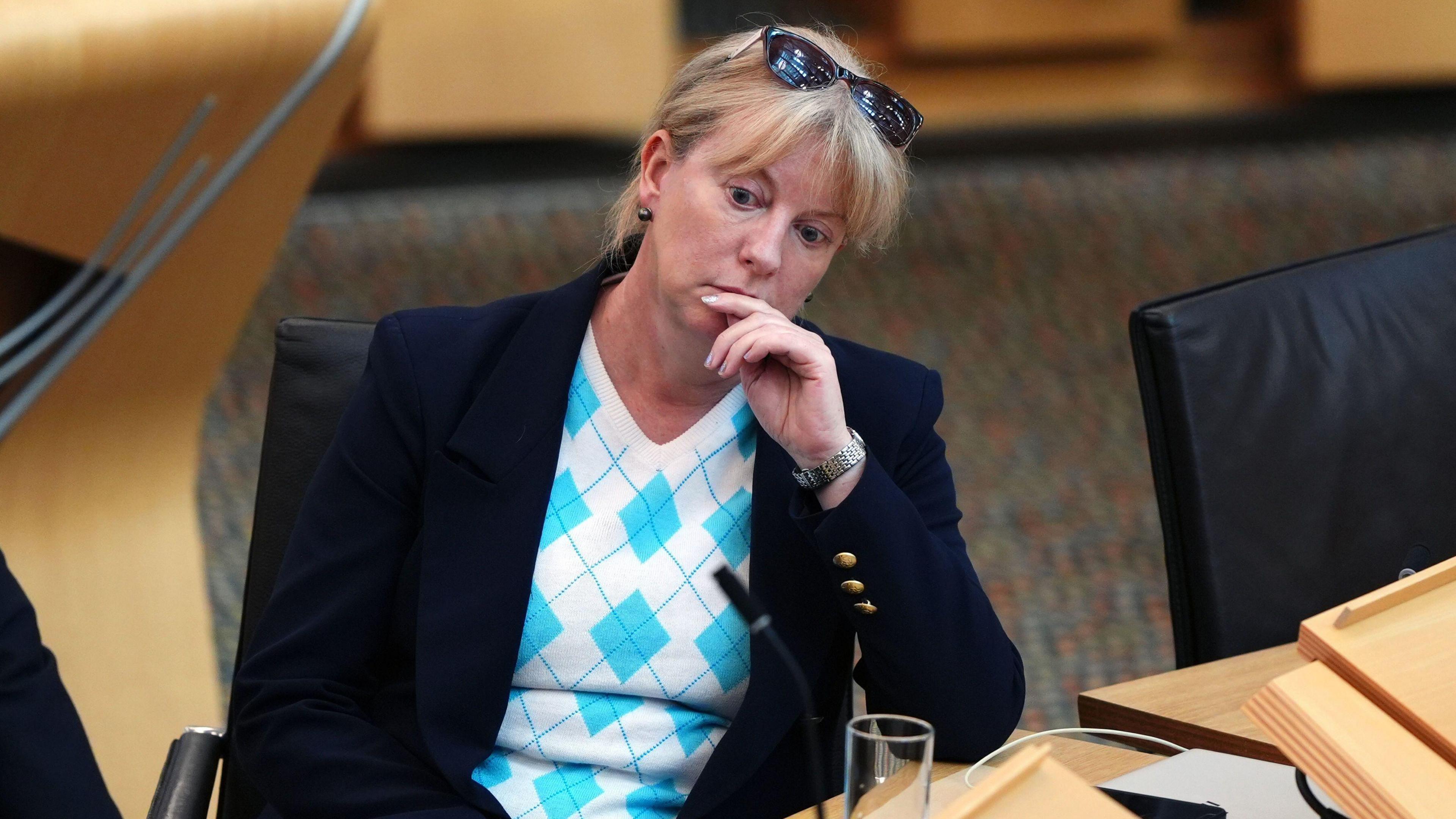 Shona Robison - a woman wearing a dark jacket and blue and white top - sits frowning in Holyrood. She has a pair of sunglasses on top of her head.