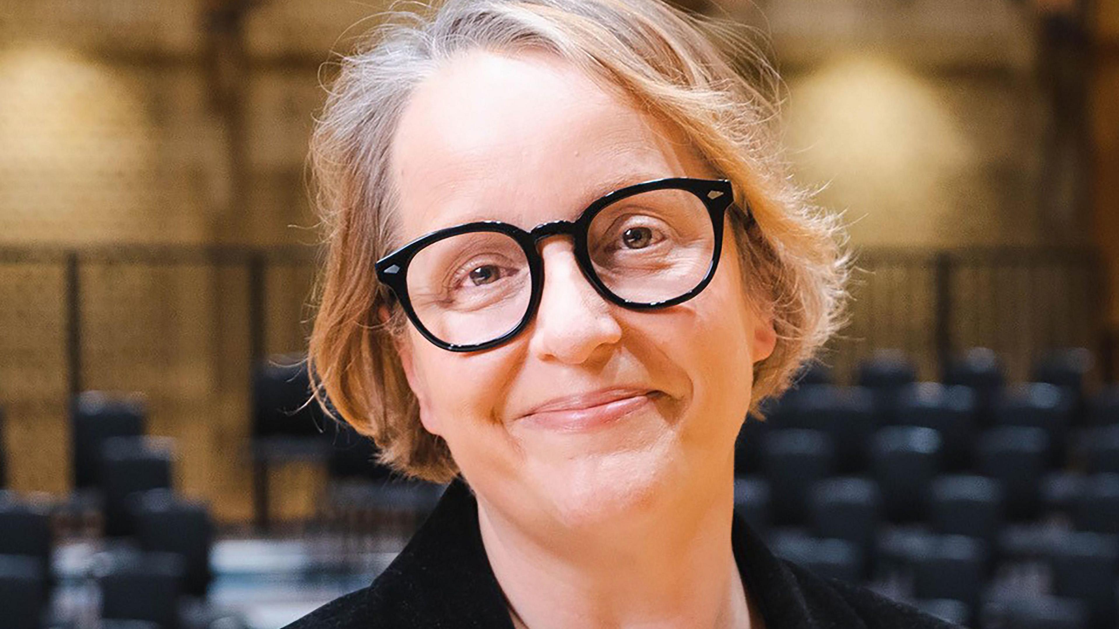 A woman with short blonde hair and black rimmed glasses, wearing a black top in a room with rows of black seats and a brown wall