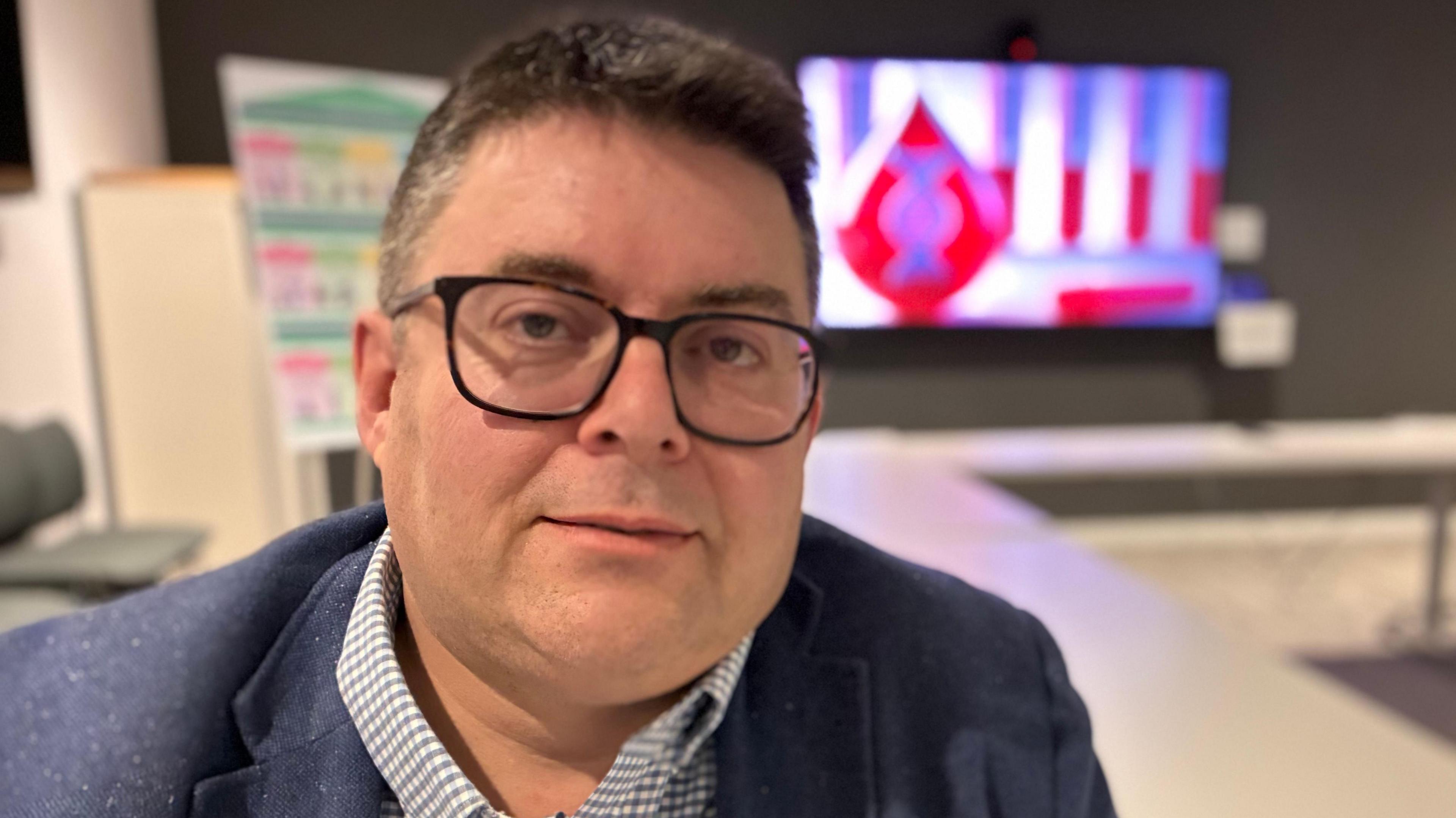 Rhodri Griffiths yn edrych at y camera. Mae'n esitedd mewn stafell cyfarfod gyda sgrin mawr yn y cefndir. Mae'n gwisgo sbectolau a jaced siwt