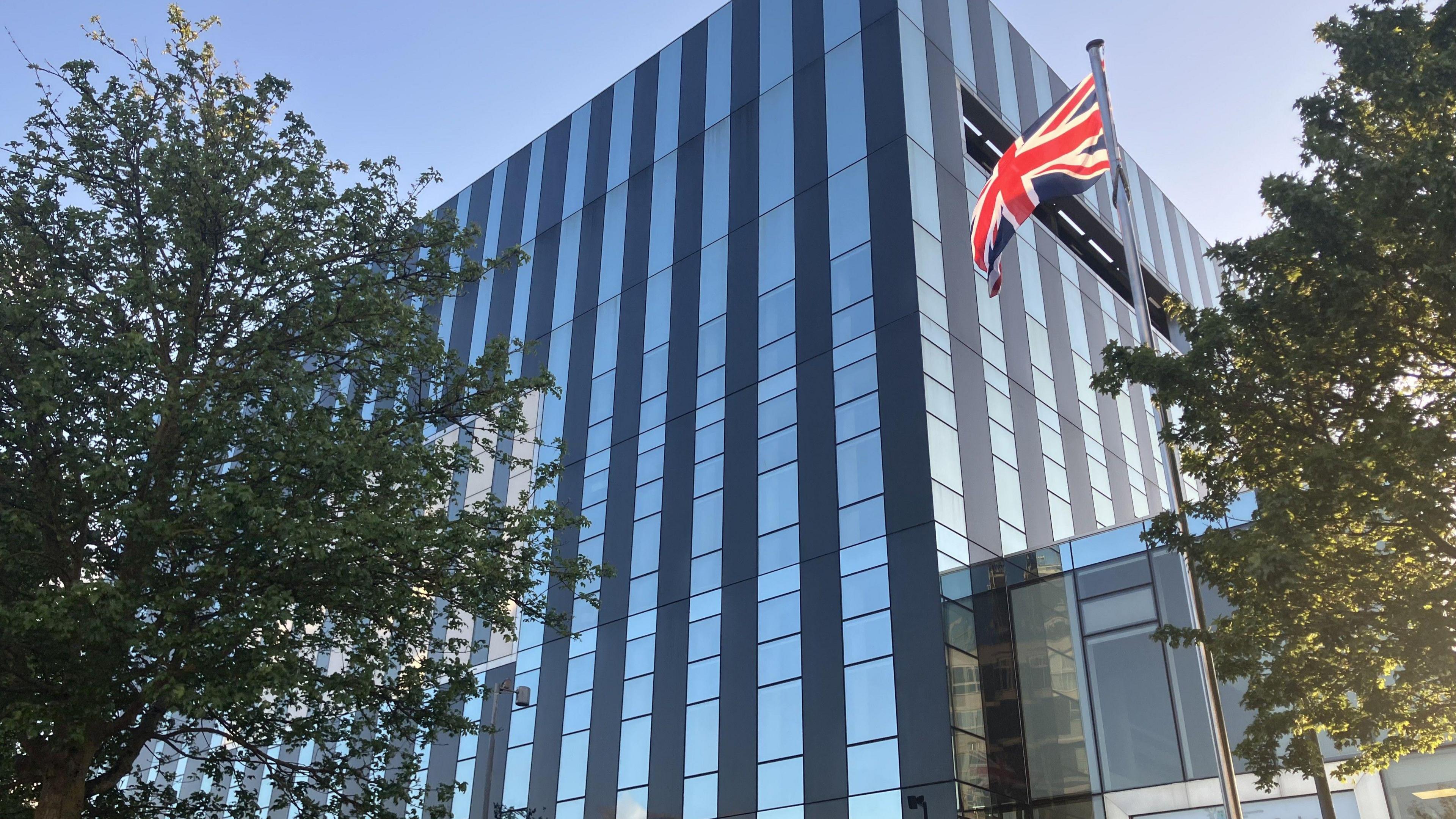 A union flag on a flagpole outside a large glass cube building. There are two trees, one on each side of the building.