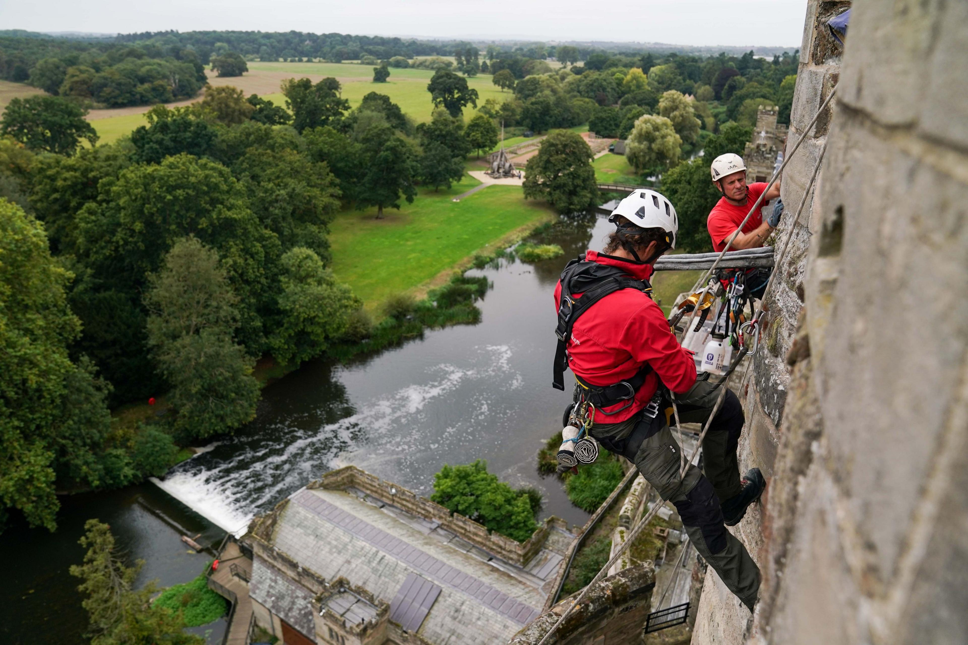 Two people in red tshirts and white helmets are abseiling stone castle walls with a view of trees and fields in the background