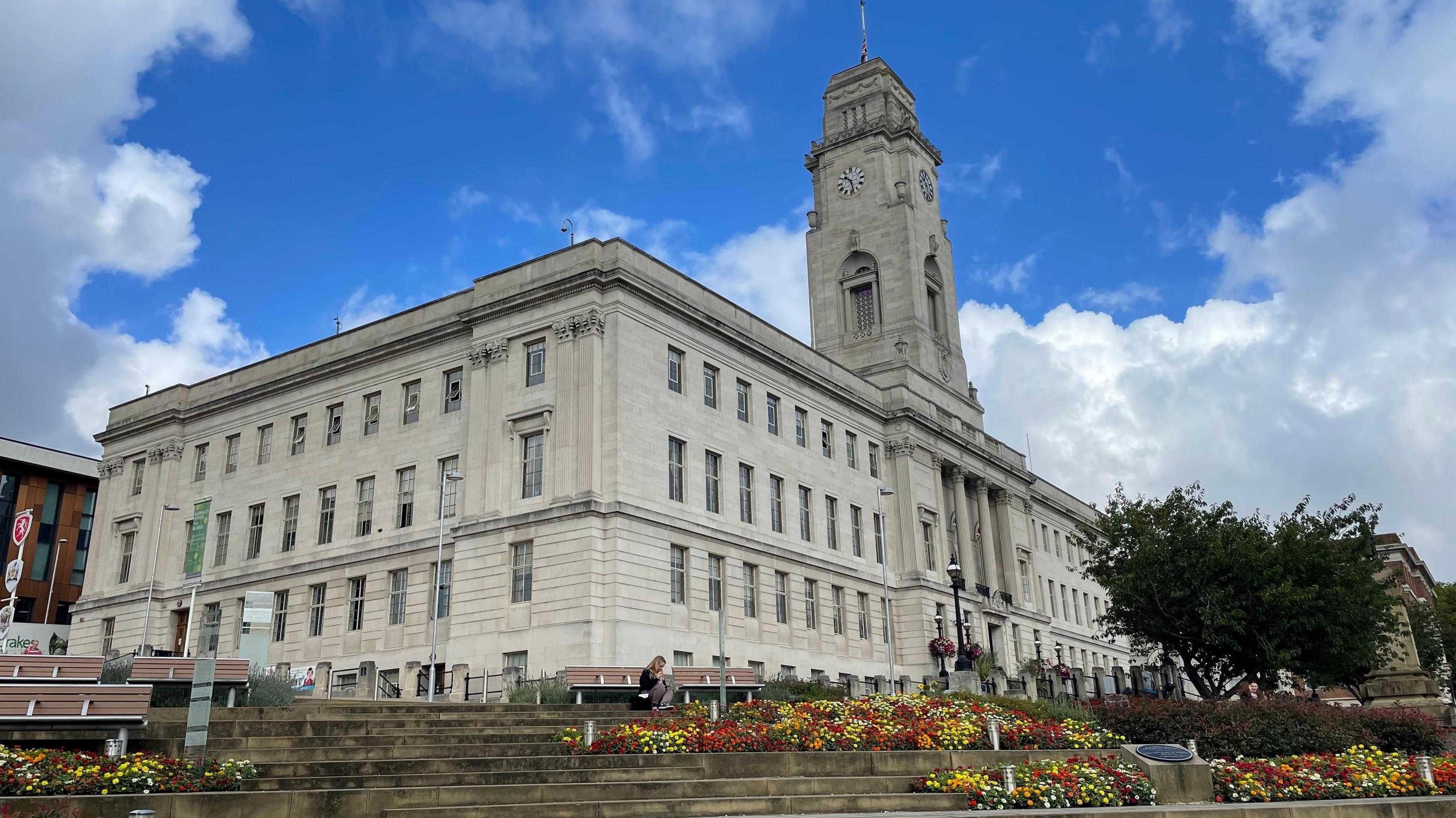 A picture of Barnsley Town Hall which is a large grey building with several floors and a clock tower. There are steps leading up the building with flowers on display. 