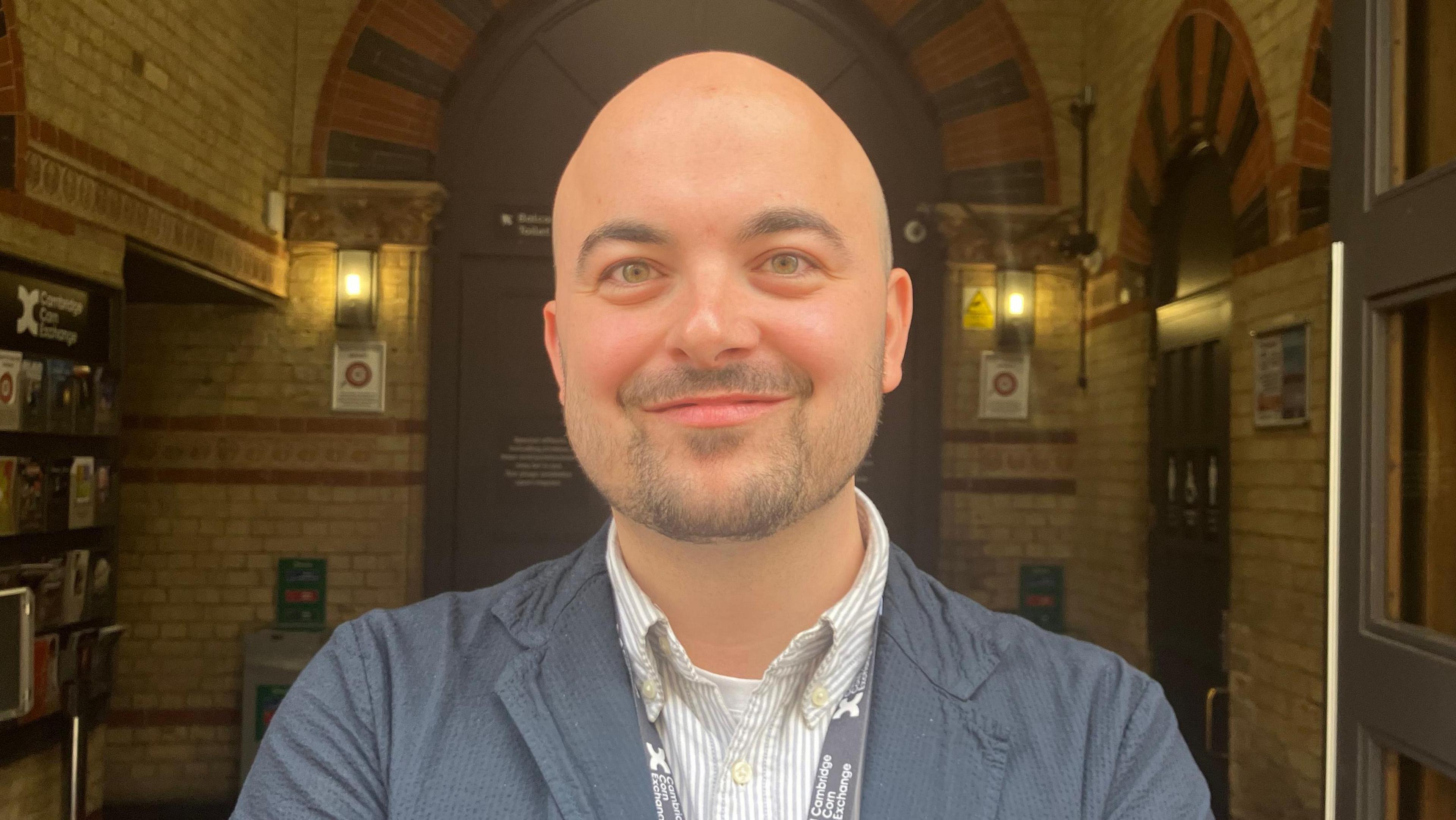 Tom Thurbon stands in front anarched doorway framed by exposed brickwork. He wears a blue jacket layered over a striped shirt, with a lanyard around his neck indicating his professional role. The background includes wall-mounted lights, signage, and an automatic door to the right.