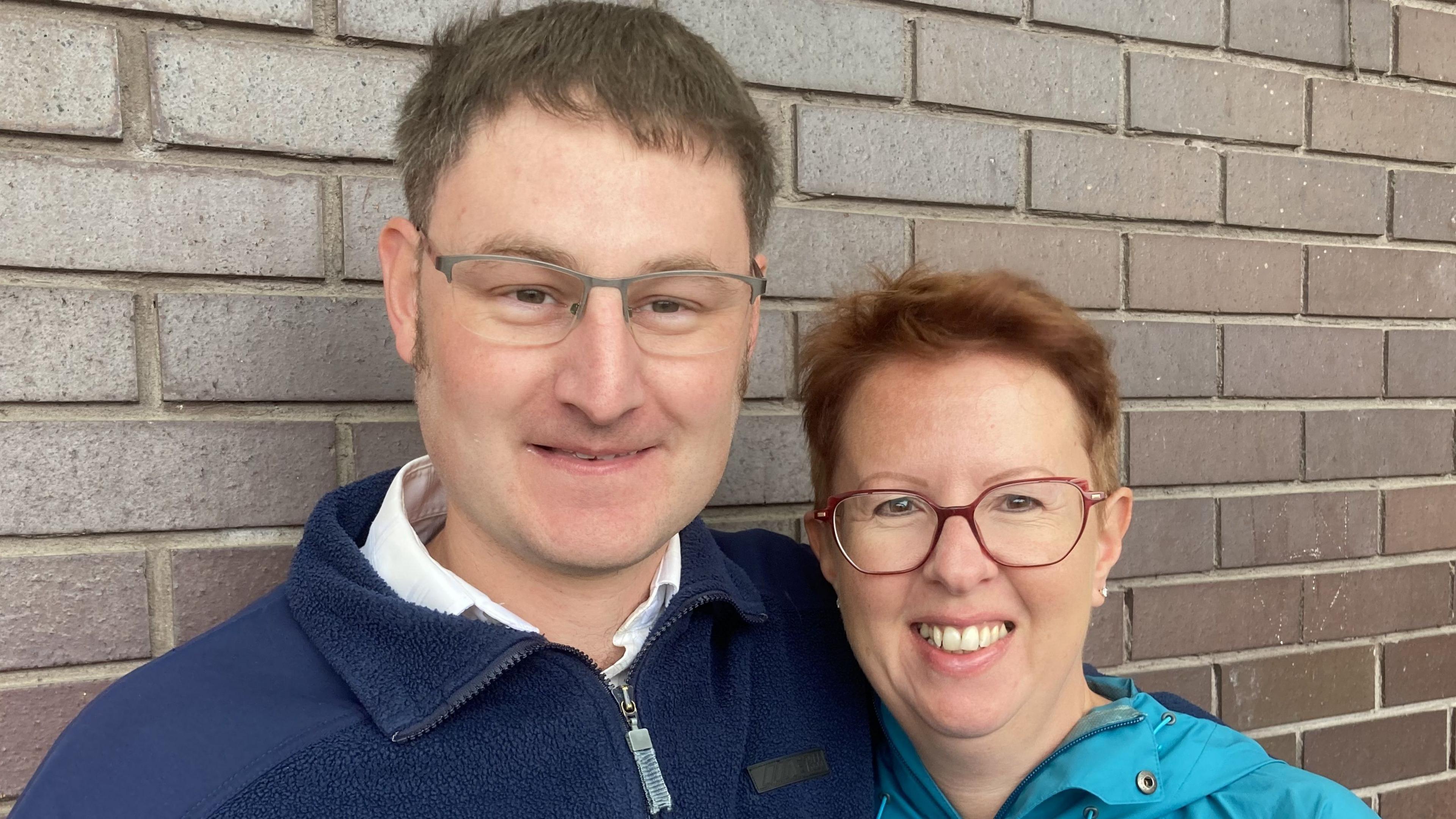 Richard Foggin and Tracy Hanlon from Swindon smiling both wearing glasses stood in front of a brown brick wall at the current bus station in Swindon.
