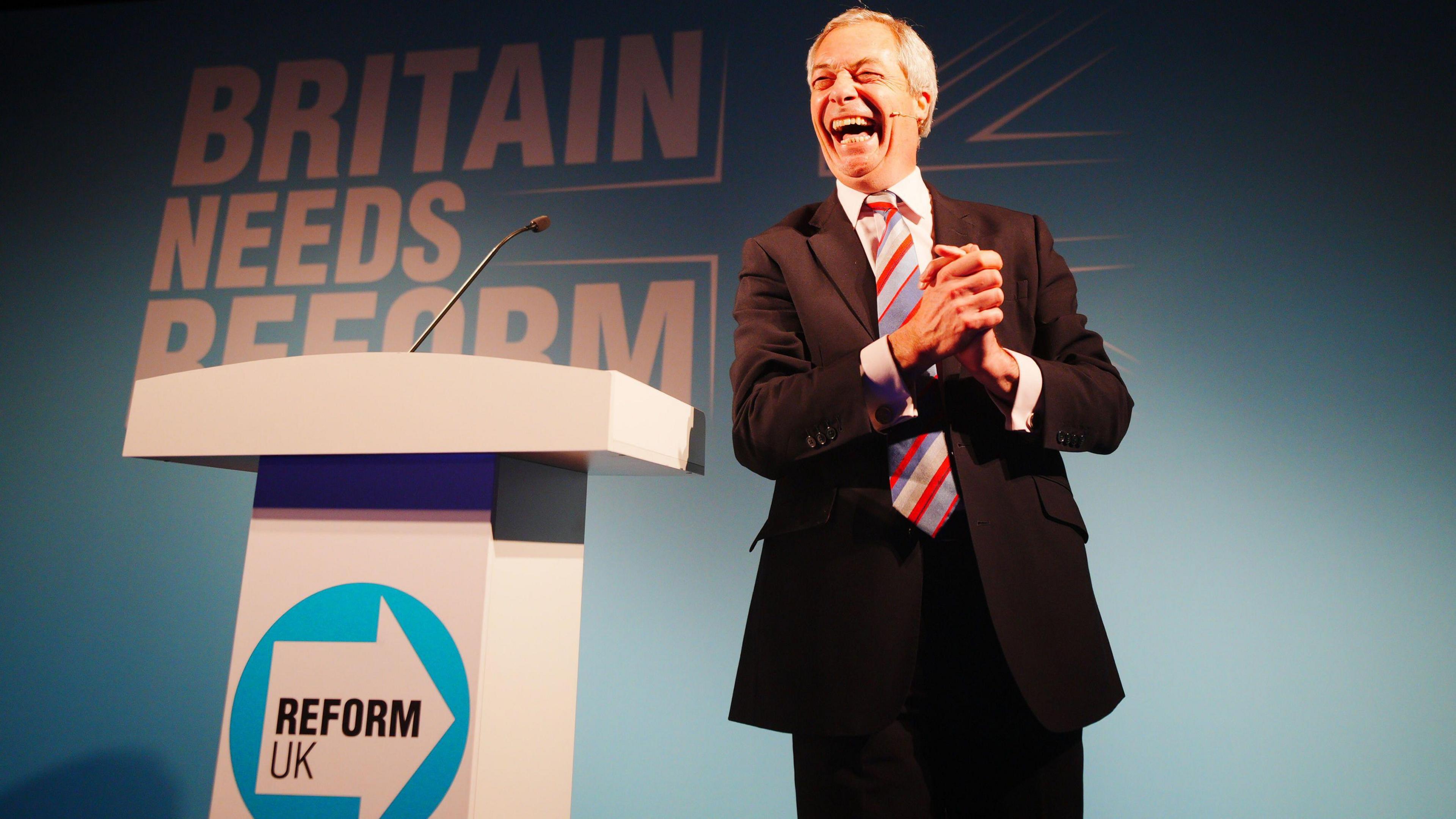 Reform UK leader Nigel Farage stands on stage in Cornwall at a rally next to a white podium with the party symbol on it. He is smiling with his mouth wide open and is stood in front of a backboard which says "Britain Needs Reform".