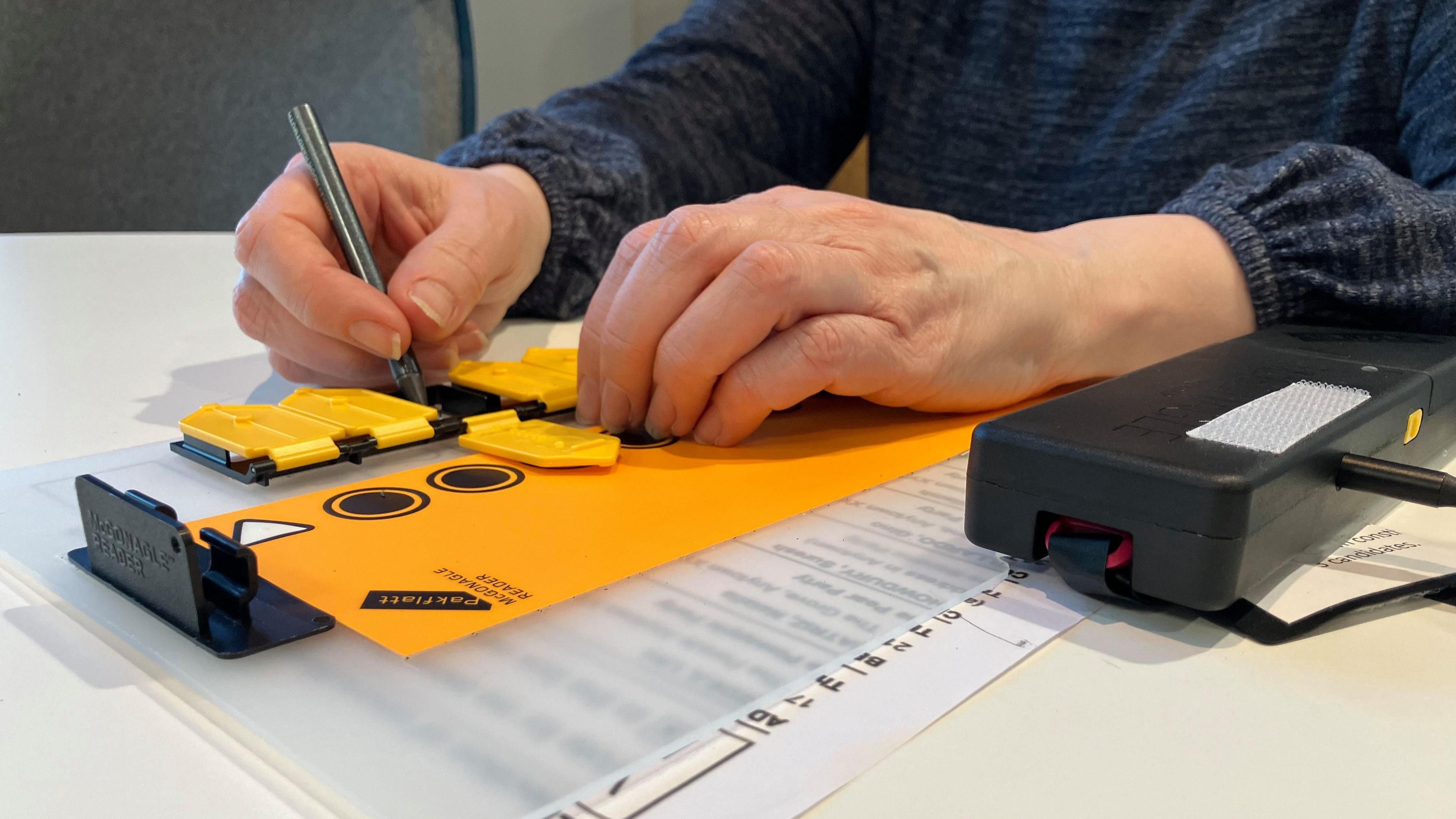 An audio-tactile device to help blind and partially-sighted people vote is on a table, placed over a ballot paper. It consists of black and yellow plastic with tabs that sit over the boxes in which voters may draw a cross. One of the tabs is open, allowing a person, whose hands are visible, to mark a cross in the box below with a pencil.