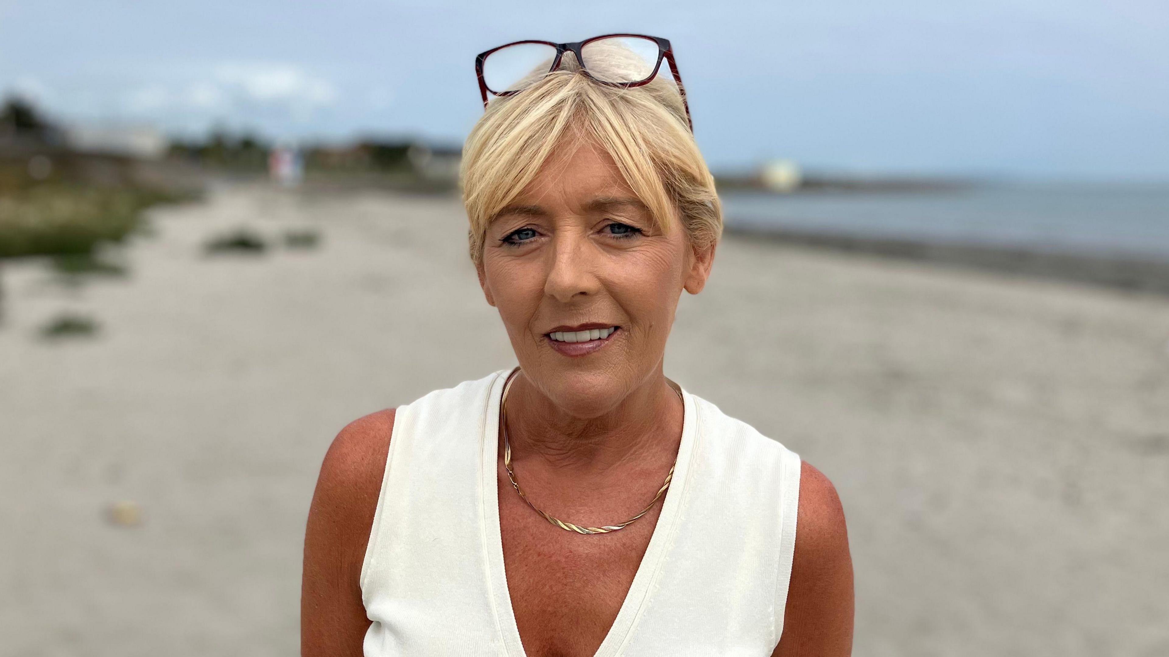 Tracy Carson has her blonde hair tied up and a pair of black-frammed glasses sitting on her head. She is wearing a white vest top and a gold necklace. She is smiling at the camera, with Millisle Beach blurred behind her.