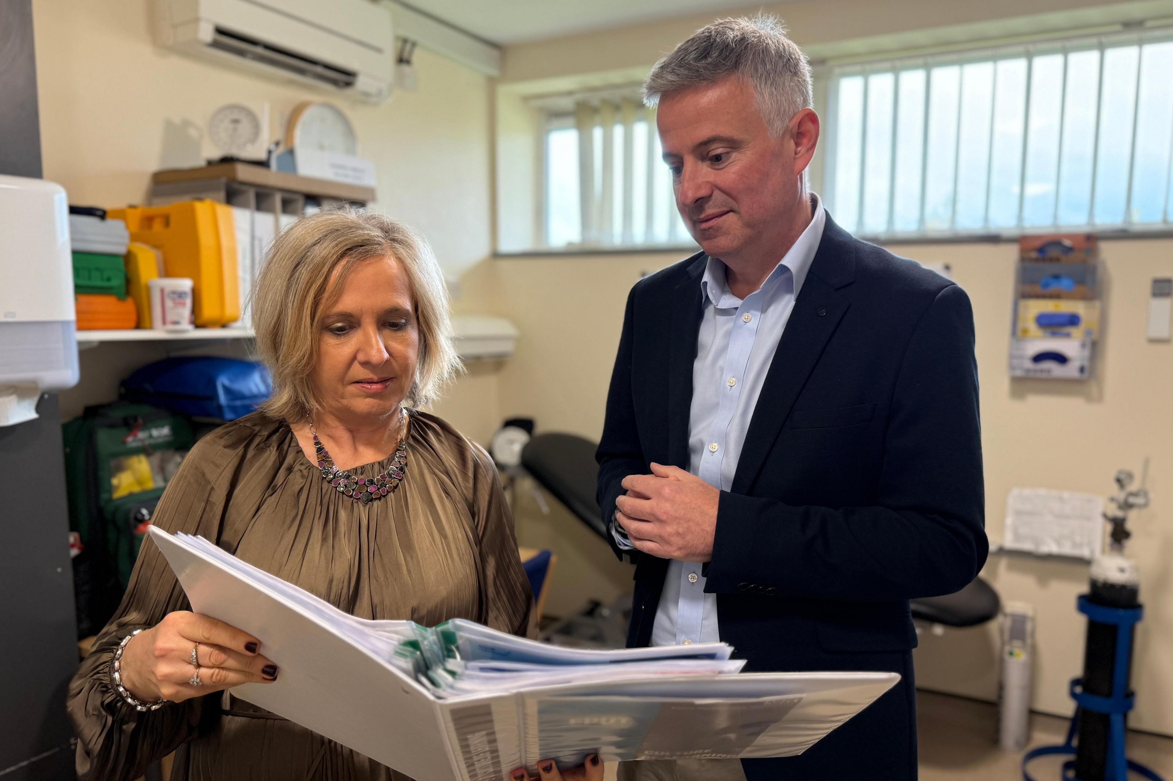 Paul Scott standing beside Martine Munby at Brockfield House. He is standing in the office with a medical bed in the background. Ms Munby is holding a white folder in front of her and is looking at it. Mr Scott is looking over Martine's shoulder a the contents of the folder. There is a high window and you can see an air conditioning unit, a clock and shelves in the background