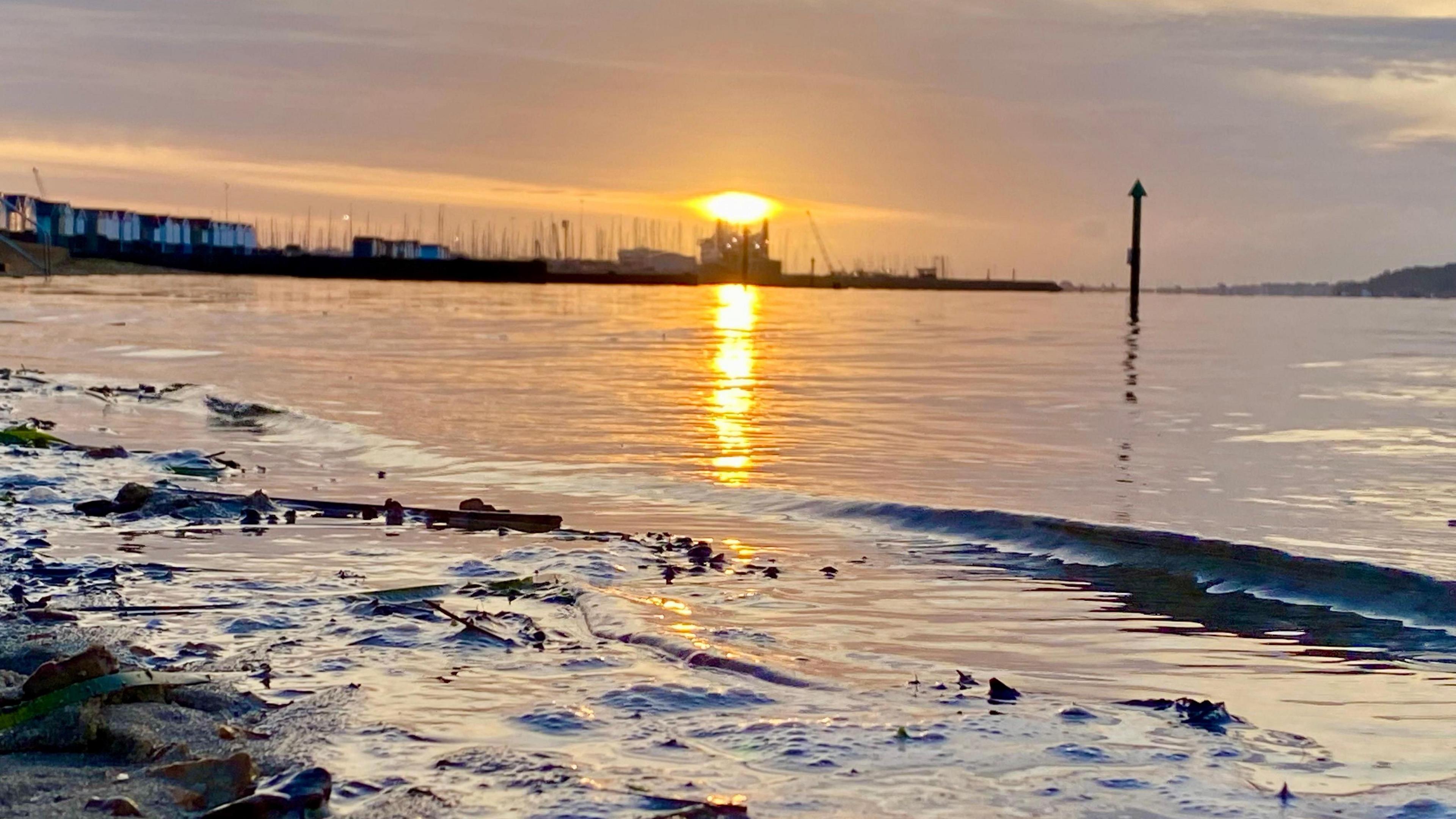 A low sun reflects into the calm water of Poole Harbour with Hamworthy's beach huts visible on the horizon 