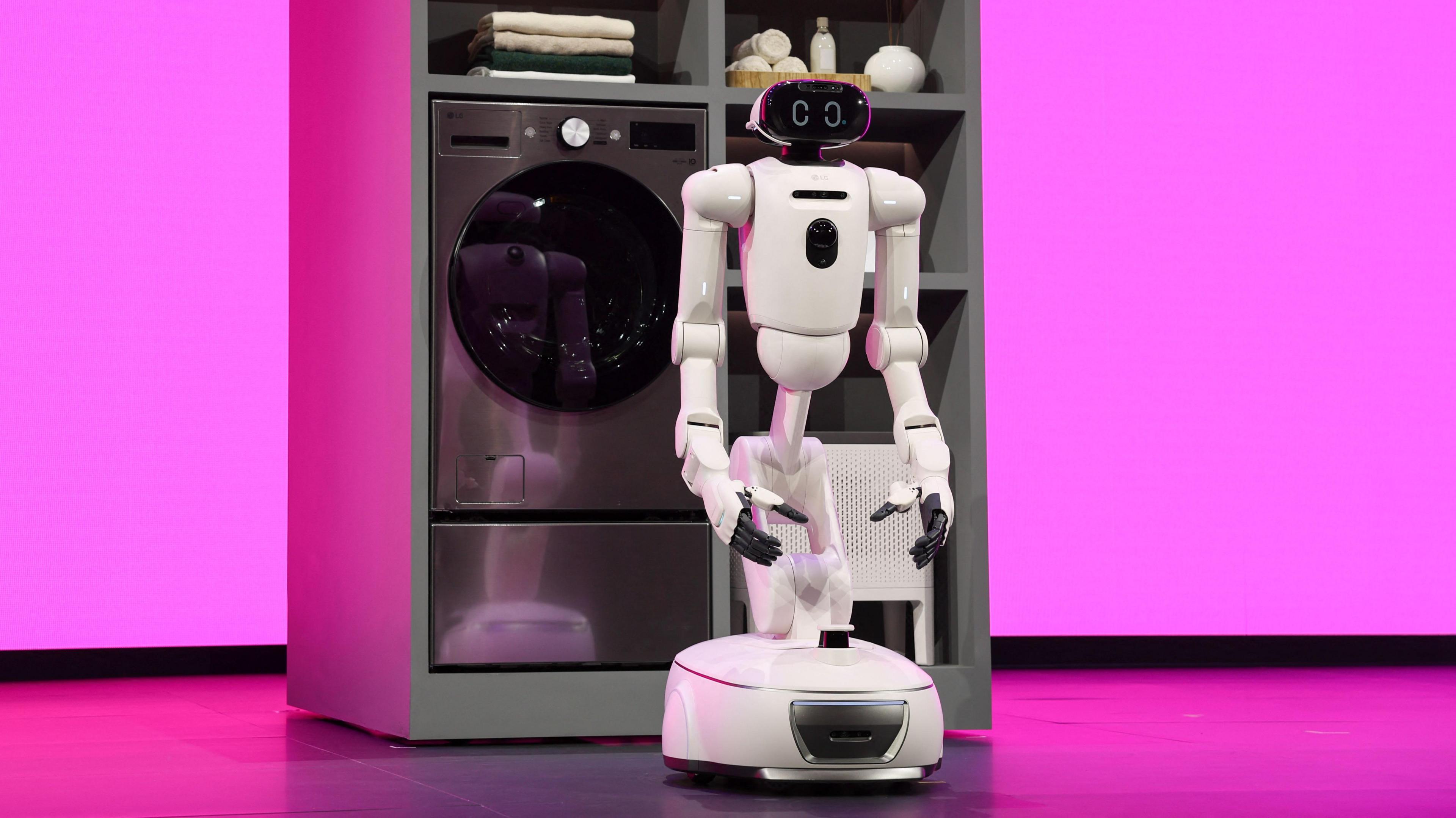 A white robot with arms and a face on a moving stand next to a dishwasher