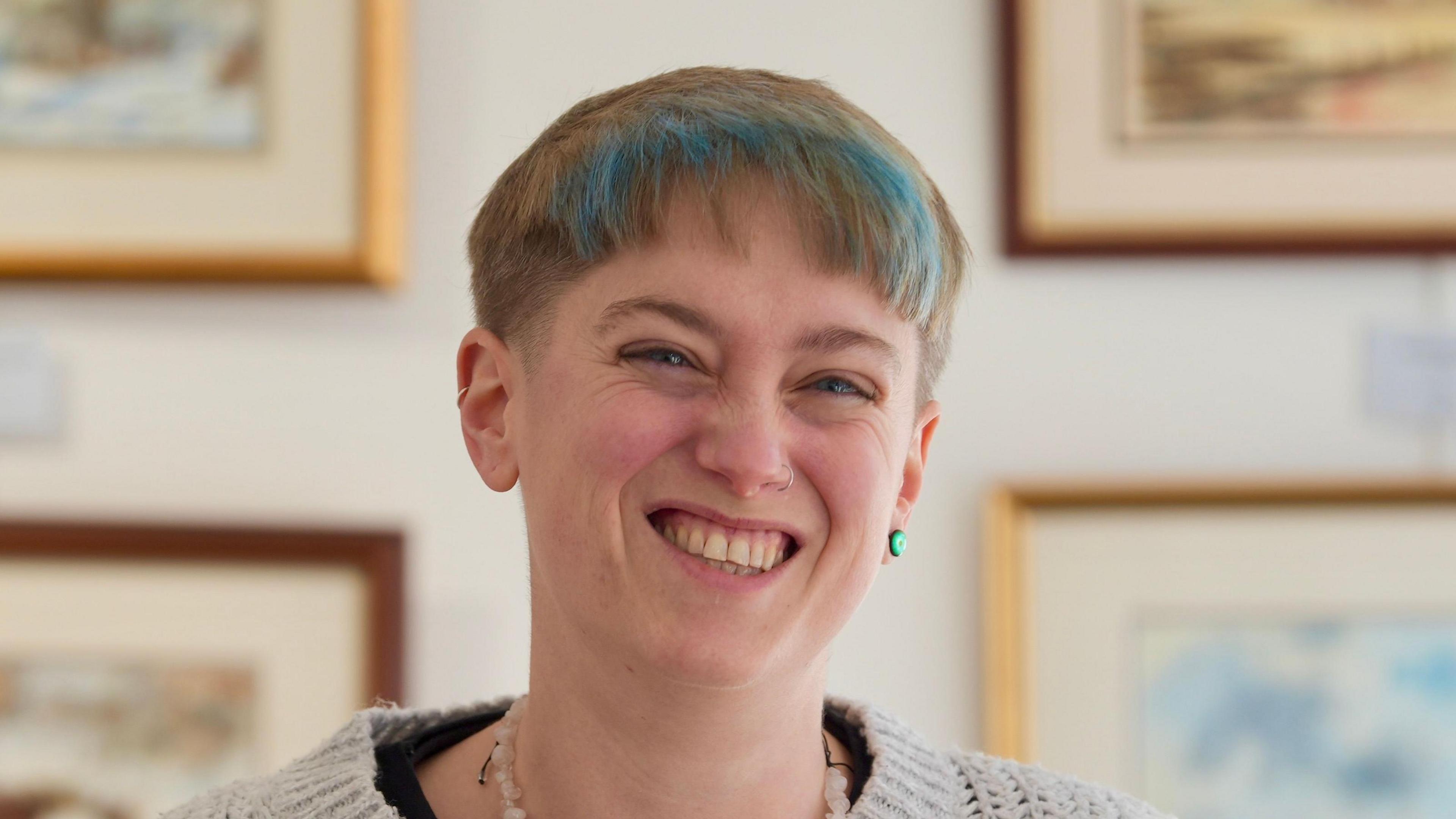 A woman with short cut strawberry blonde hair and dyed blue parts in a fringe. She is smiling and has one green earring. She is wearing a white knitted jumper. Pieces of framed artwork rest on the wall behind her.