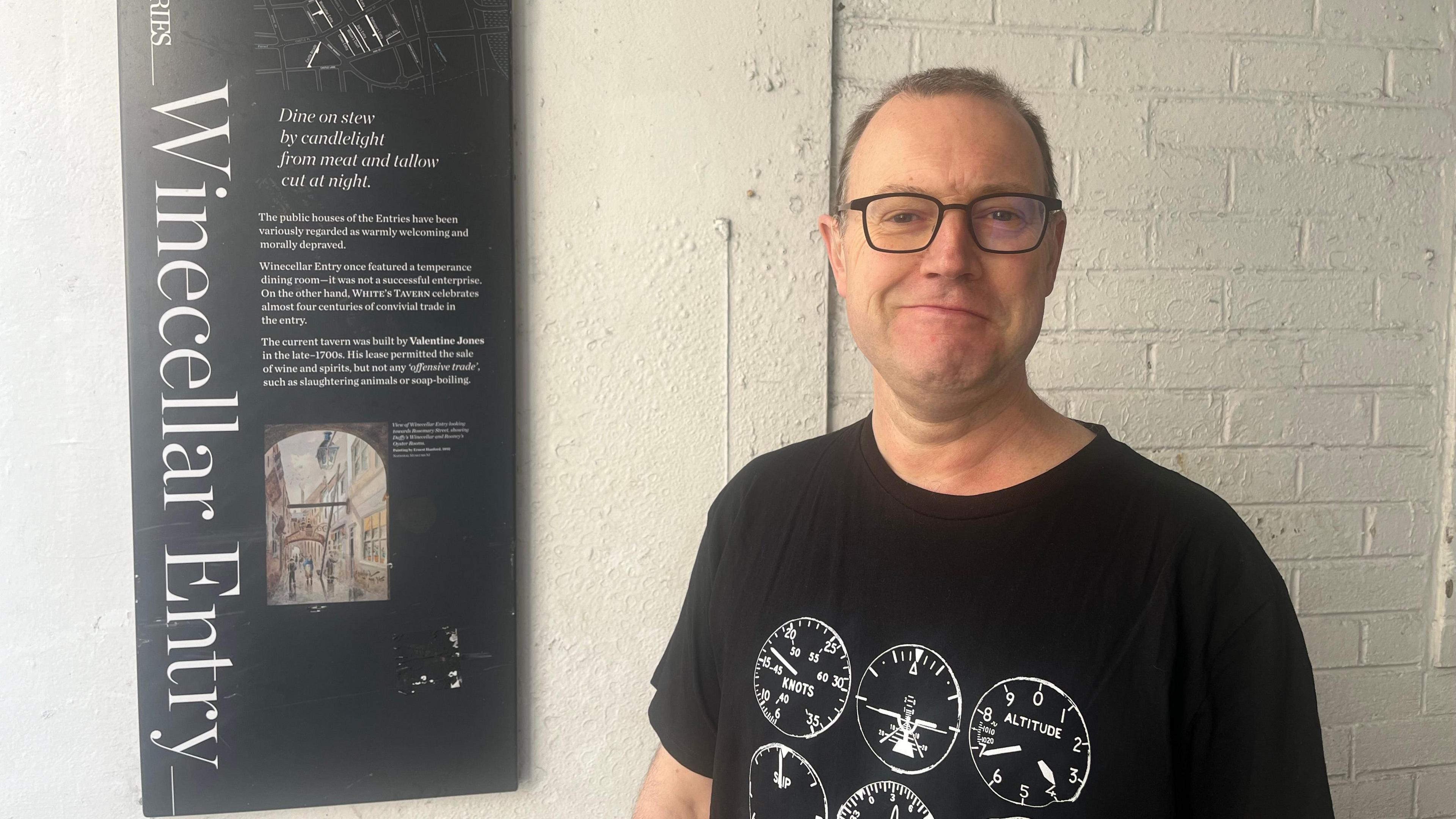 A man in a black t shirt he has brown hair and is wearing glasses. Beside him is a black sign that says winecellar entry