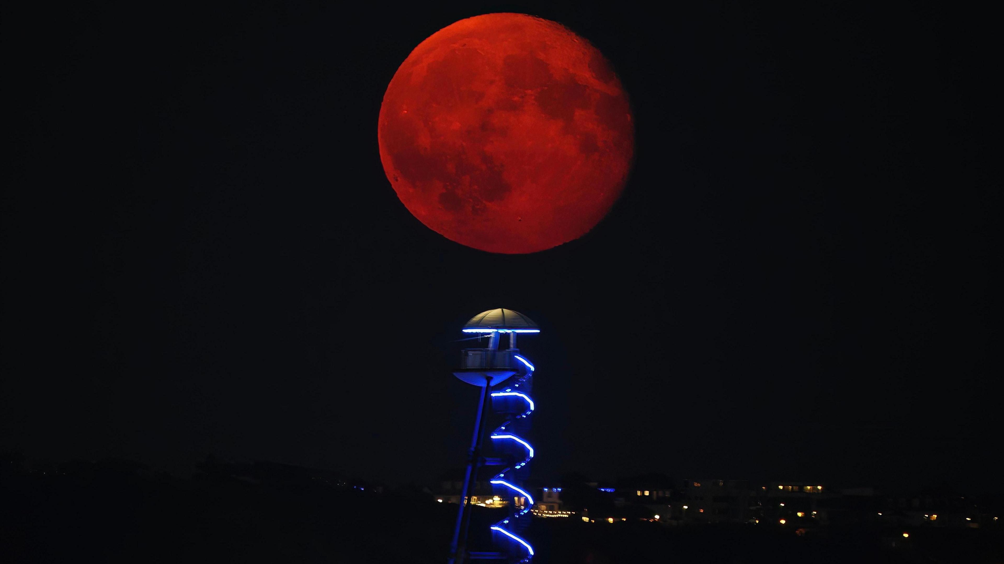 A red-looking moon can be seen above a helter skelter lit up in the dark on Bournemouth seafront. Lights from buildings can be seen in the dark background.