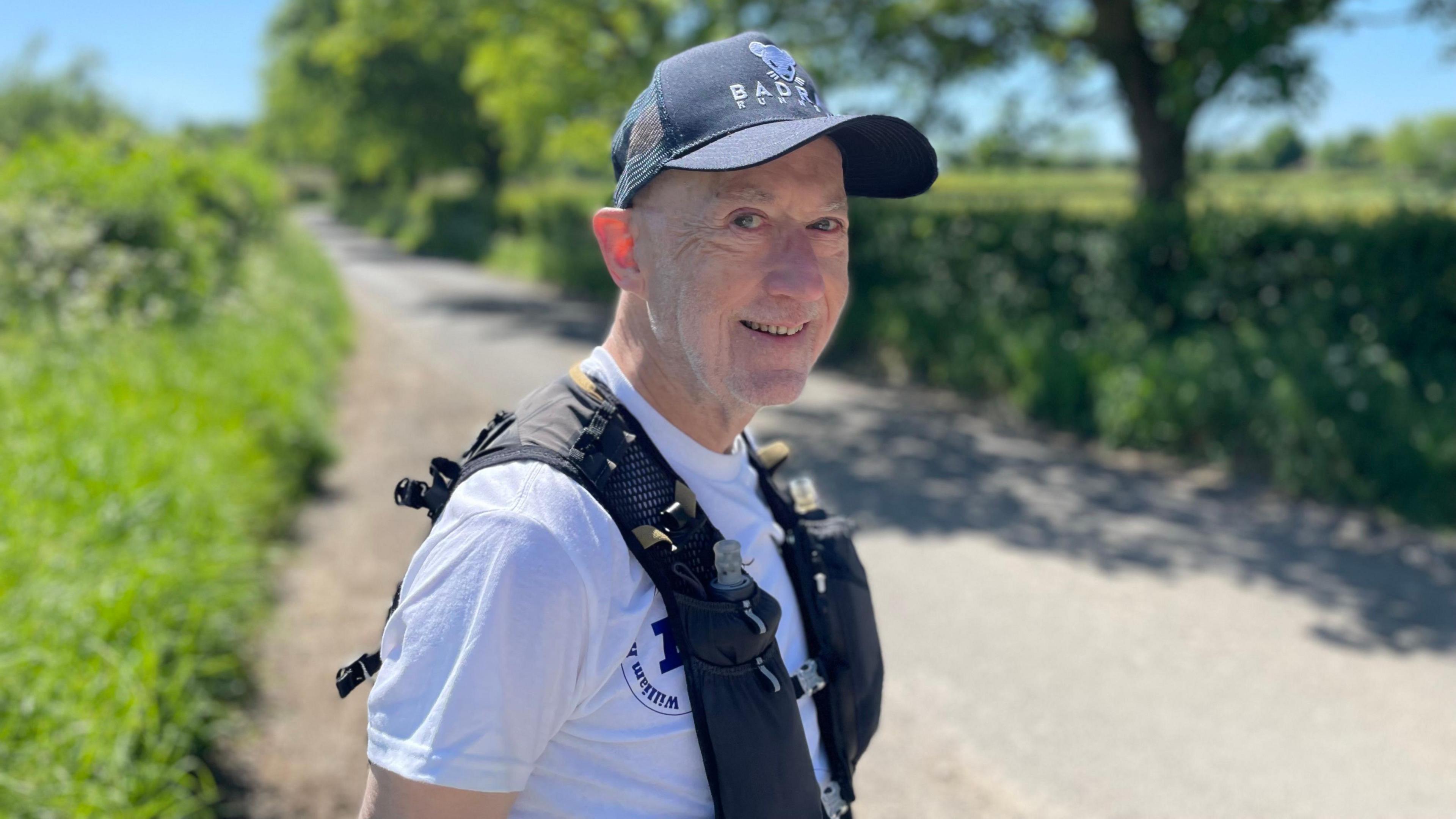 Peter smiling at the camera wearing a dark baseball cap and white T-shirt underneath a black running vest with water bottles inside its pockets. He is stood in a layby on a country lane, with blurred hedges and trees in the background.