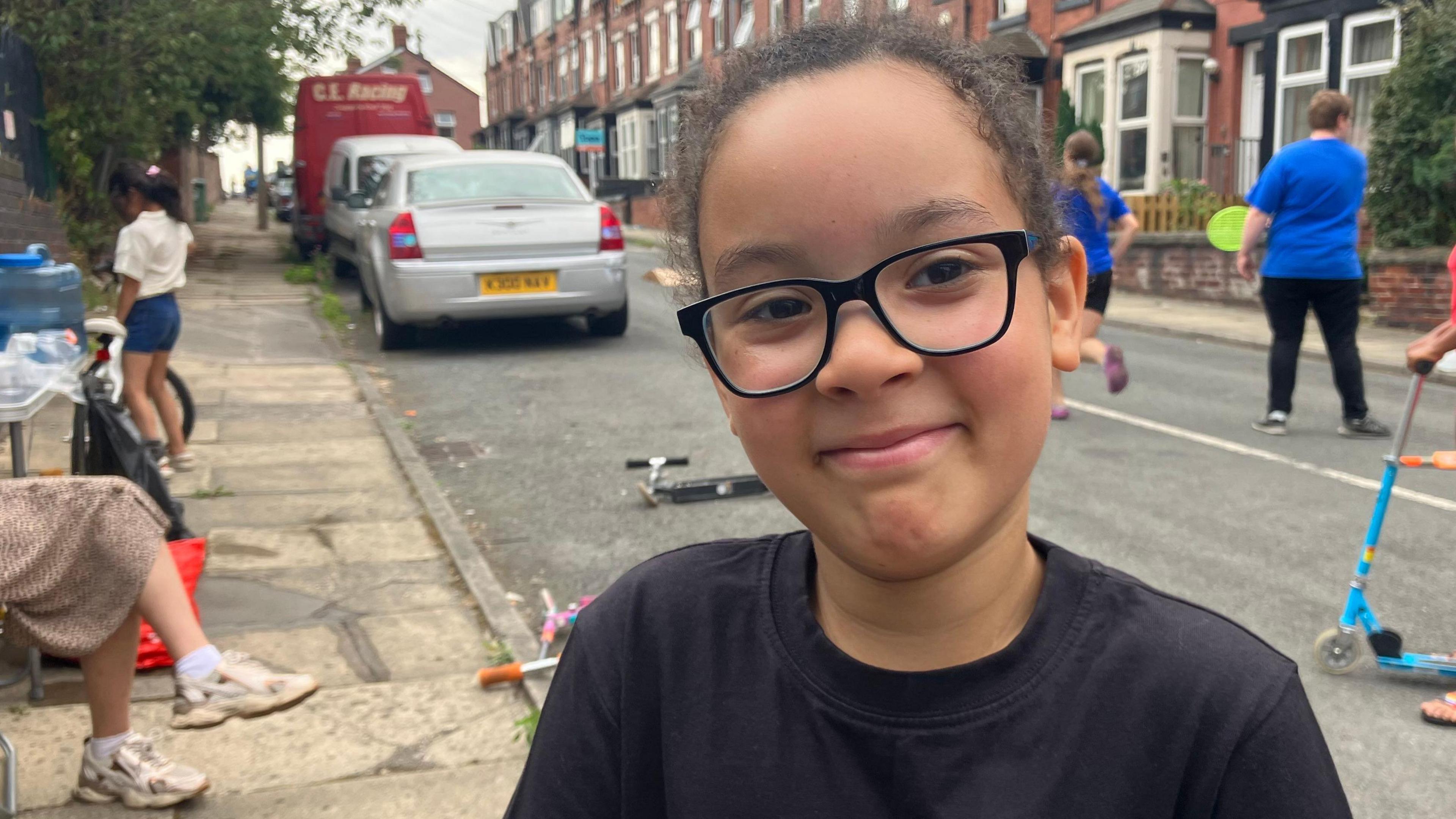 A 7 twelvemonth aged lad wearing a achromatic t garment and achromatic glasses is smiling astatine the camera successful the centre of frame. Behind him are children playing connected scooters and with tennis bats and balls successful a residential street, portion parents beryllium connected the pavement.