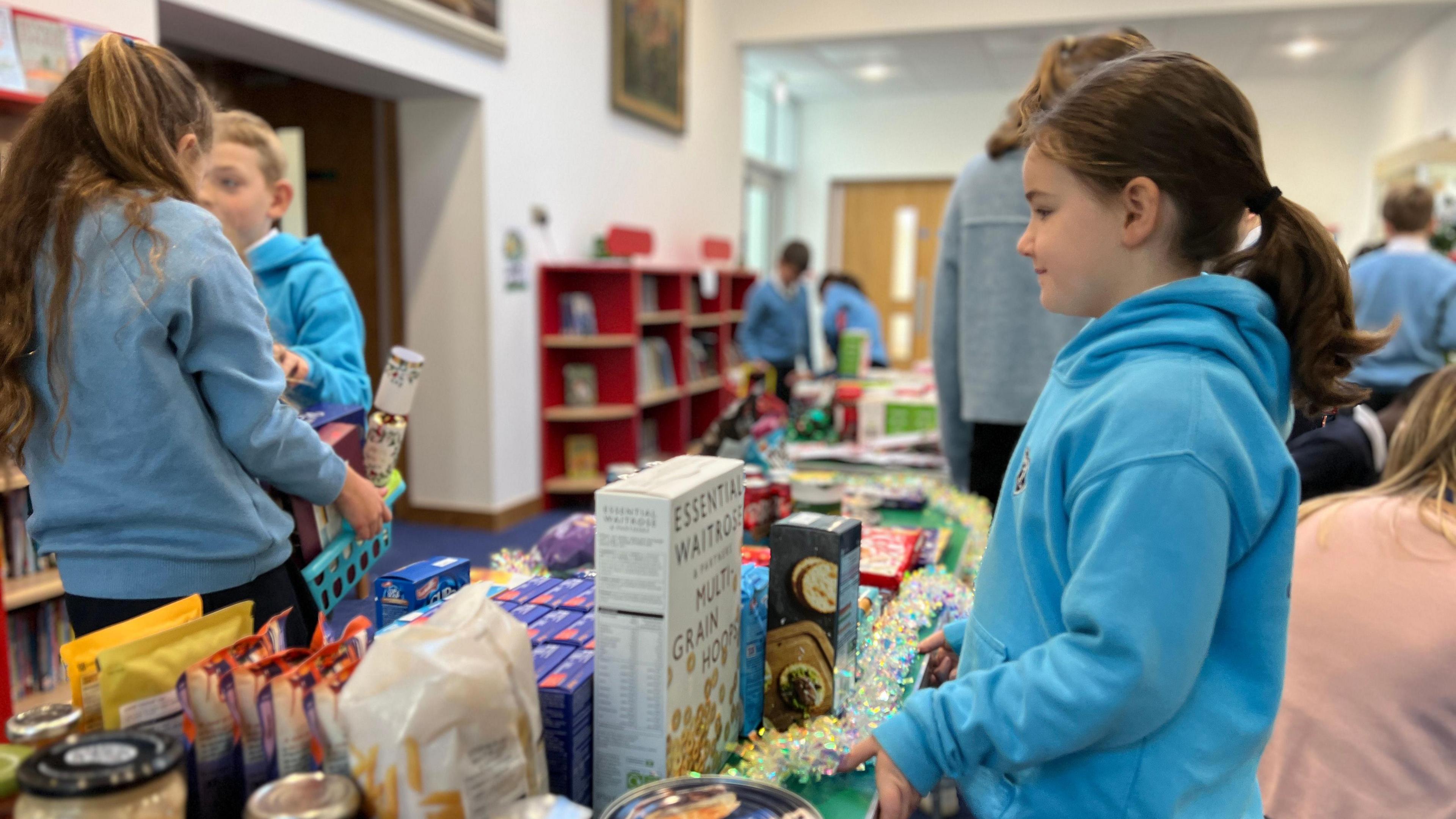 A young girl stands by a table with food packets and boxes in front of her. She has dark brown hair tied into a ponytail. Around her - other children sort through the donations and food to make the hampers.