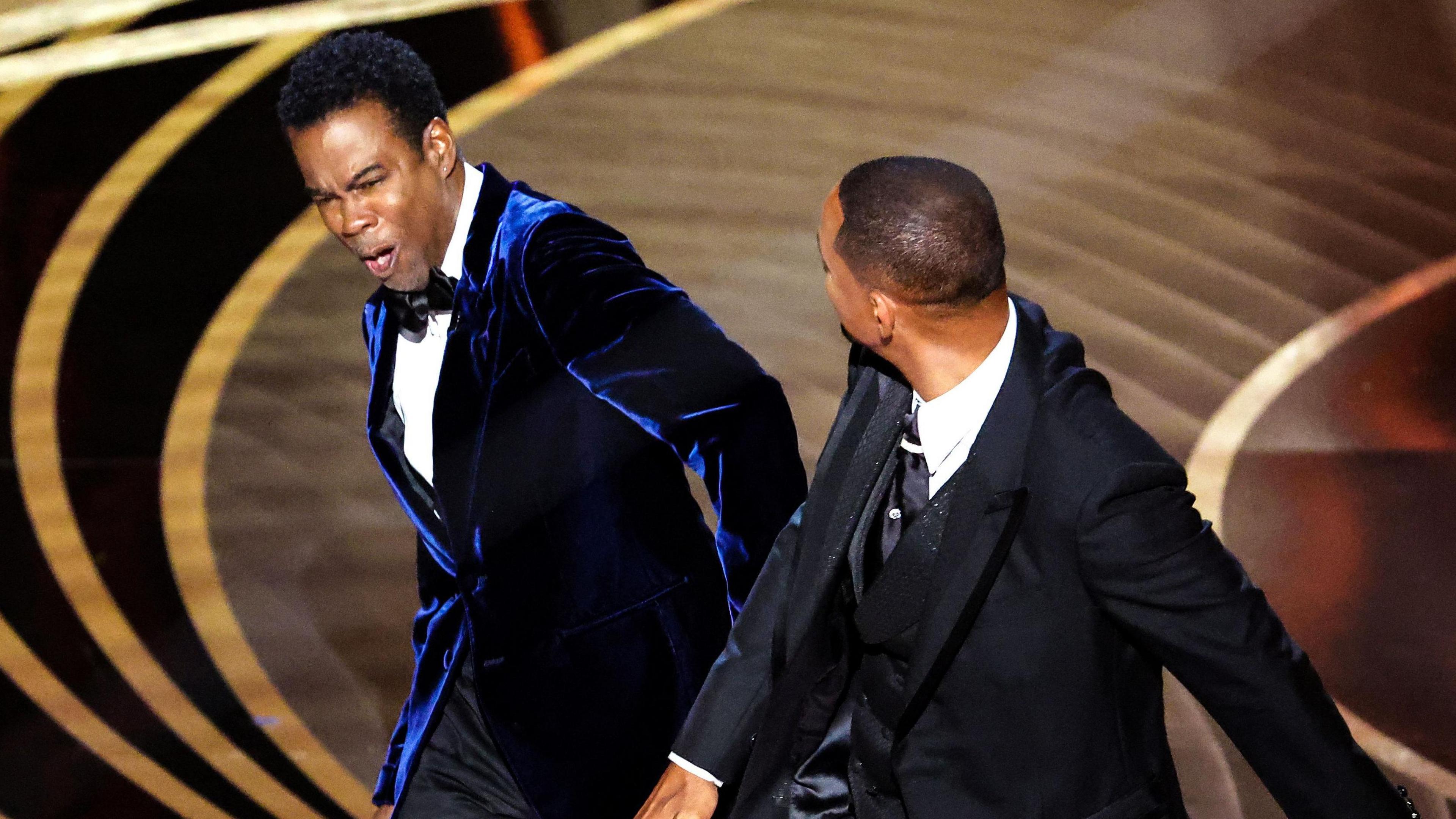 Will Smith slaps Chris Rock onstage during the show at the 94th Academy Awards at the Dolby Theatre at Ovation Hollywood on Sunday, March 27, 2022. Chris Rock, wearing a blue velvet suit and bow tie, grimaces after being struck. Will Smith, pictured from the back, wears a black tux, his right arm in front of him after striking the comedian on stage.