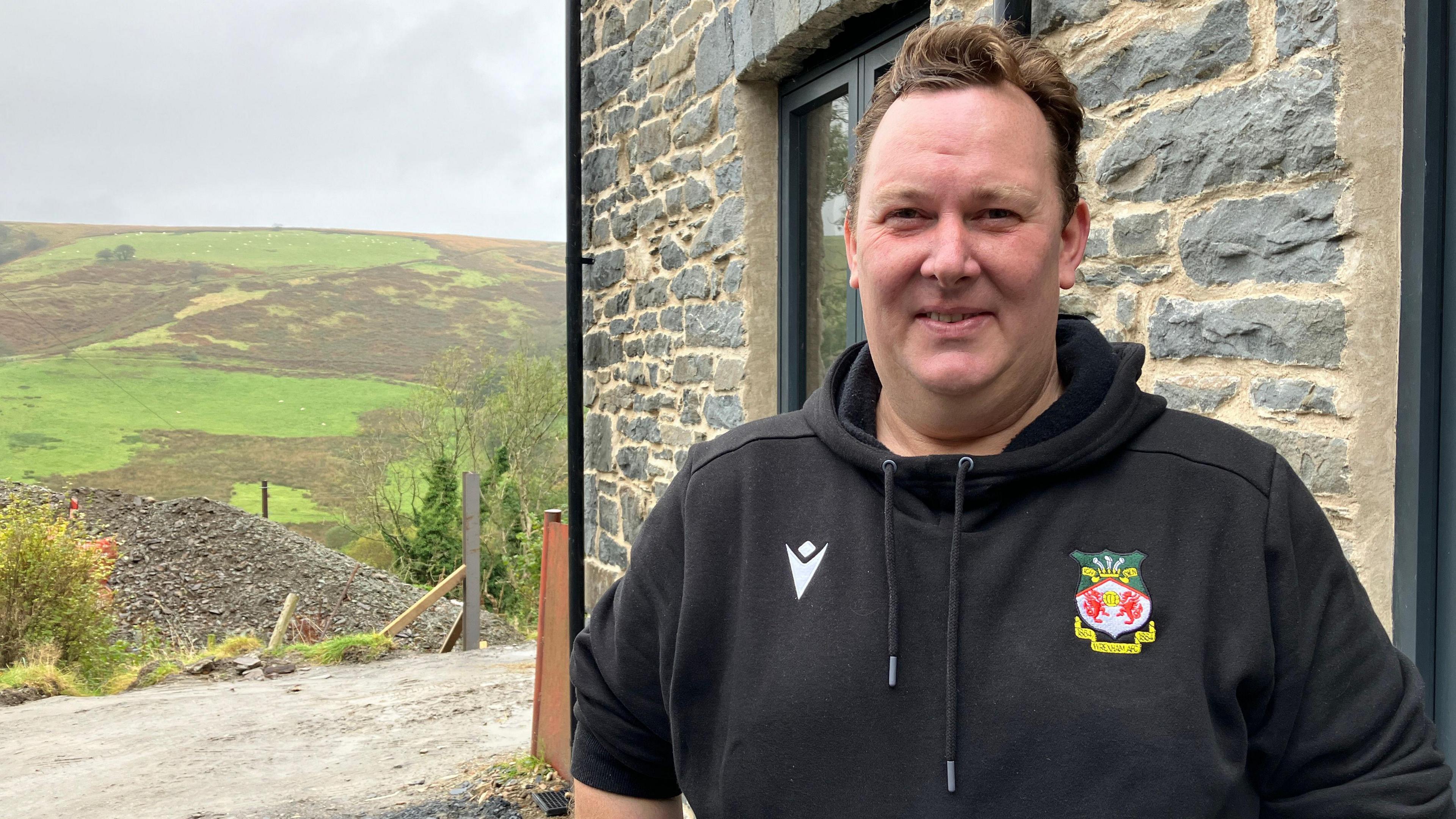 Aled Wyn Davies wearing a black hoodie and standing in front of a house with green hills in the background