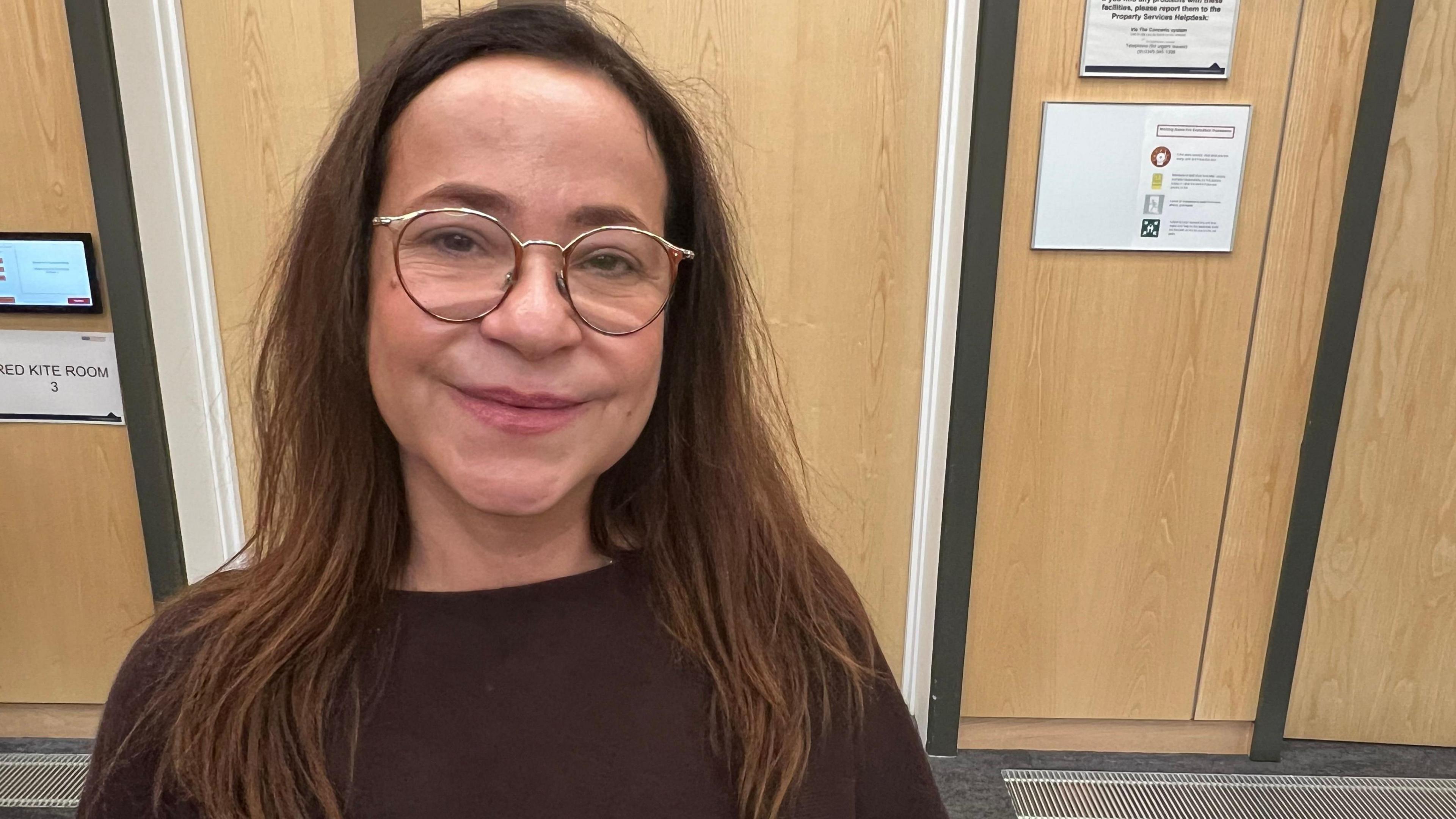 A woman with long brown hair and glasses smiles at the camera. She is wearing a brown crewneck top. Behind her is a light-coloured wooden wall and door.