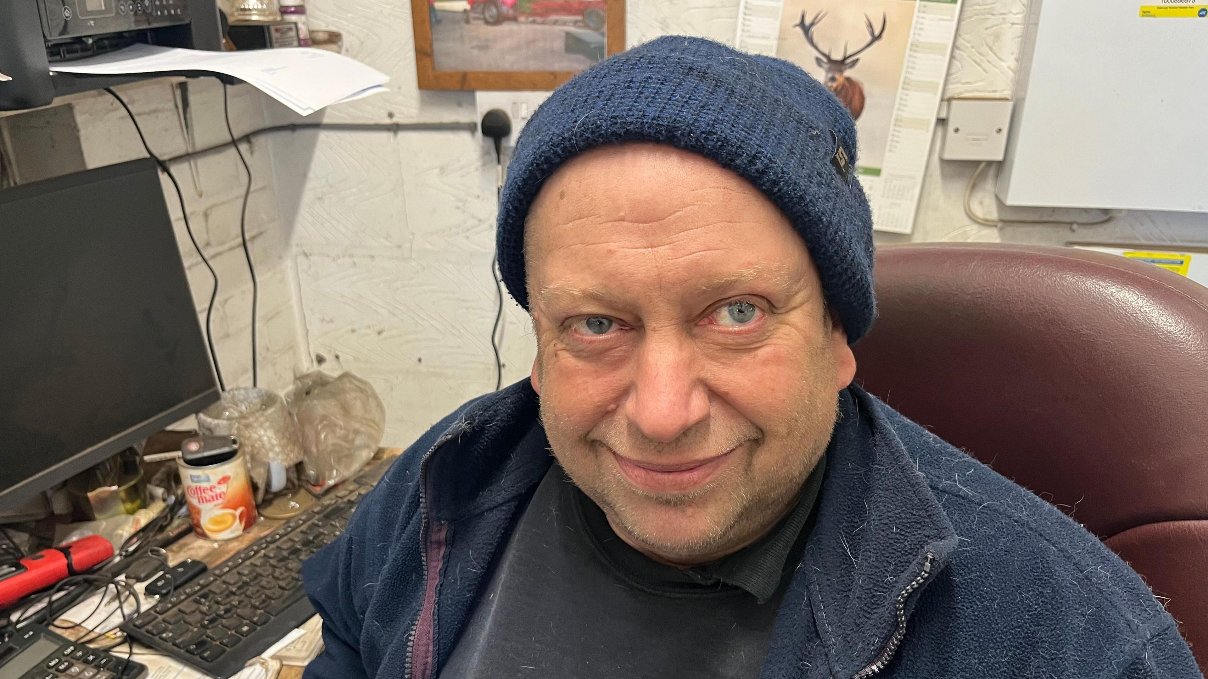 Steve Neugebauer, 55, has a blue fleece over a black jumper, he is wearing a blue beany hat and has a computer and keyboard on a desk behind him, he is sitting in a small office.