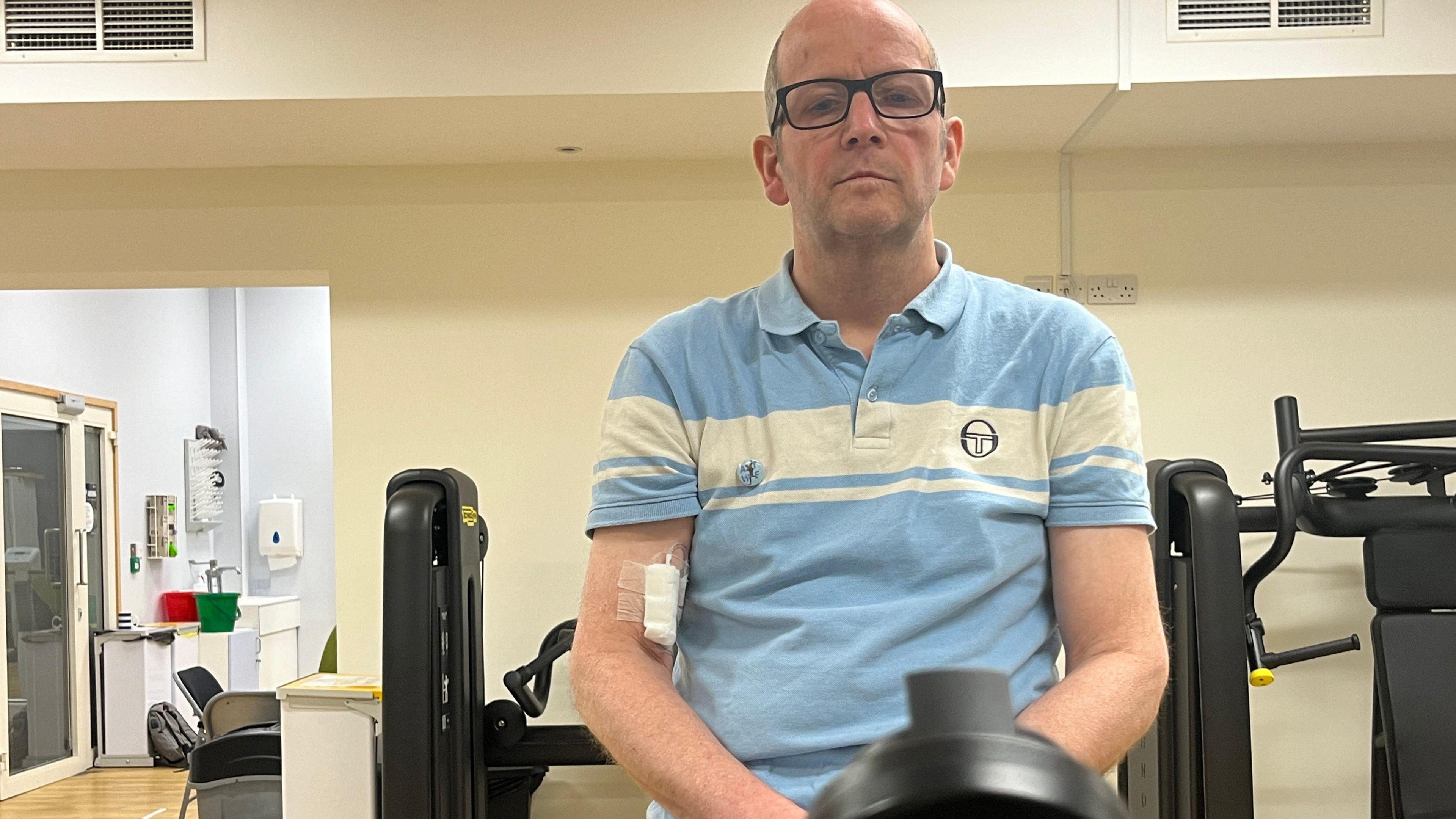 A man wearing a blue and white polo top sitting on an exercise bike in a gym. He has a piece of white bandage taped to his arm.
